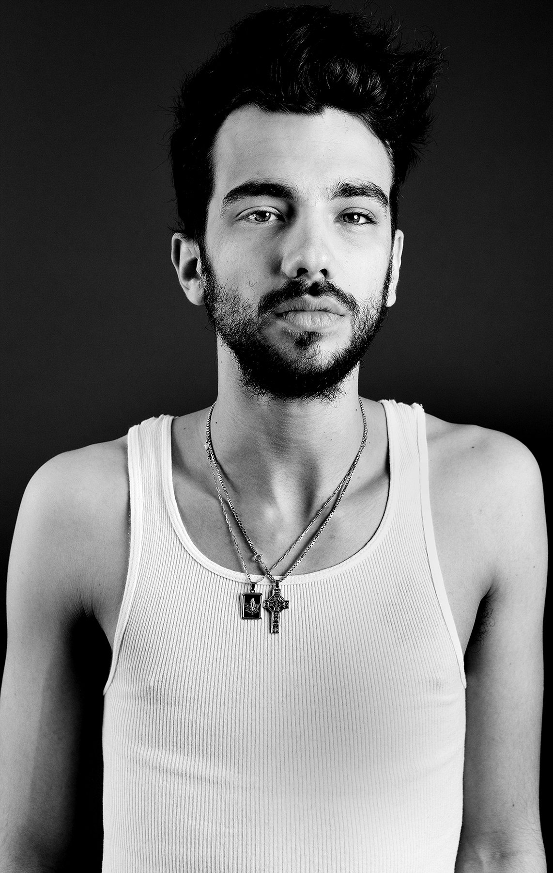 Black and white portrait of a young man with dark hair and a beard, wearing a white tank top and multiple necklaces, against a dark background.