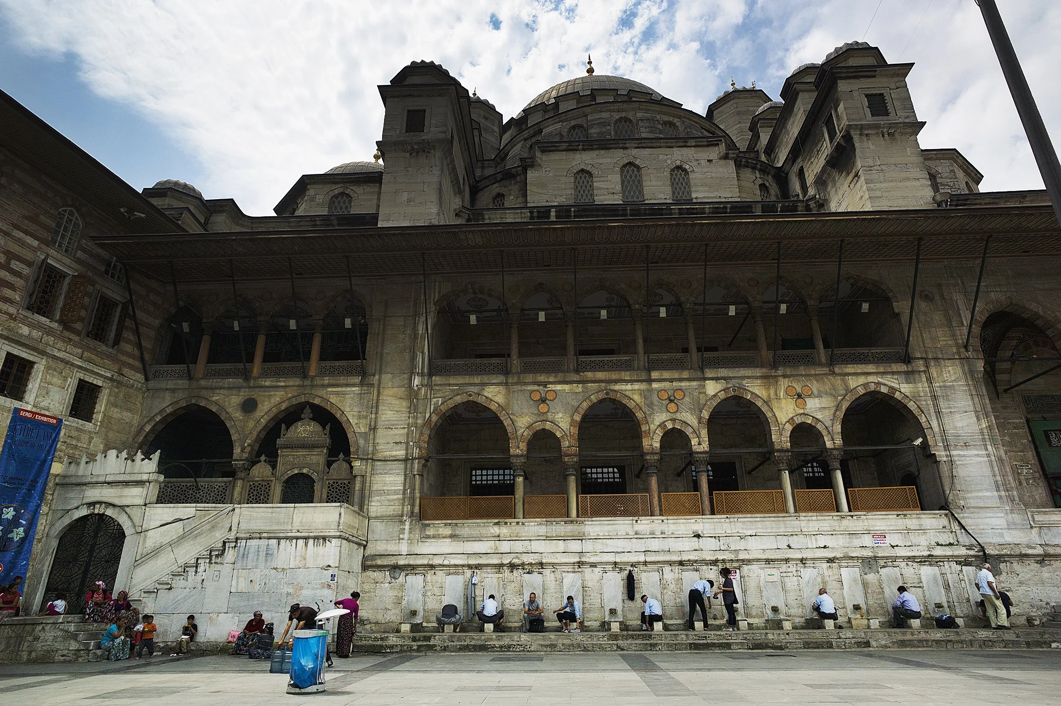 Exterior view of a historic mosque with multiple domes, arches, and intricate architecture, people sitting and standing outside in an open plaza.