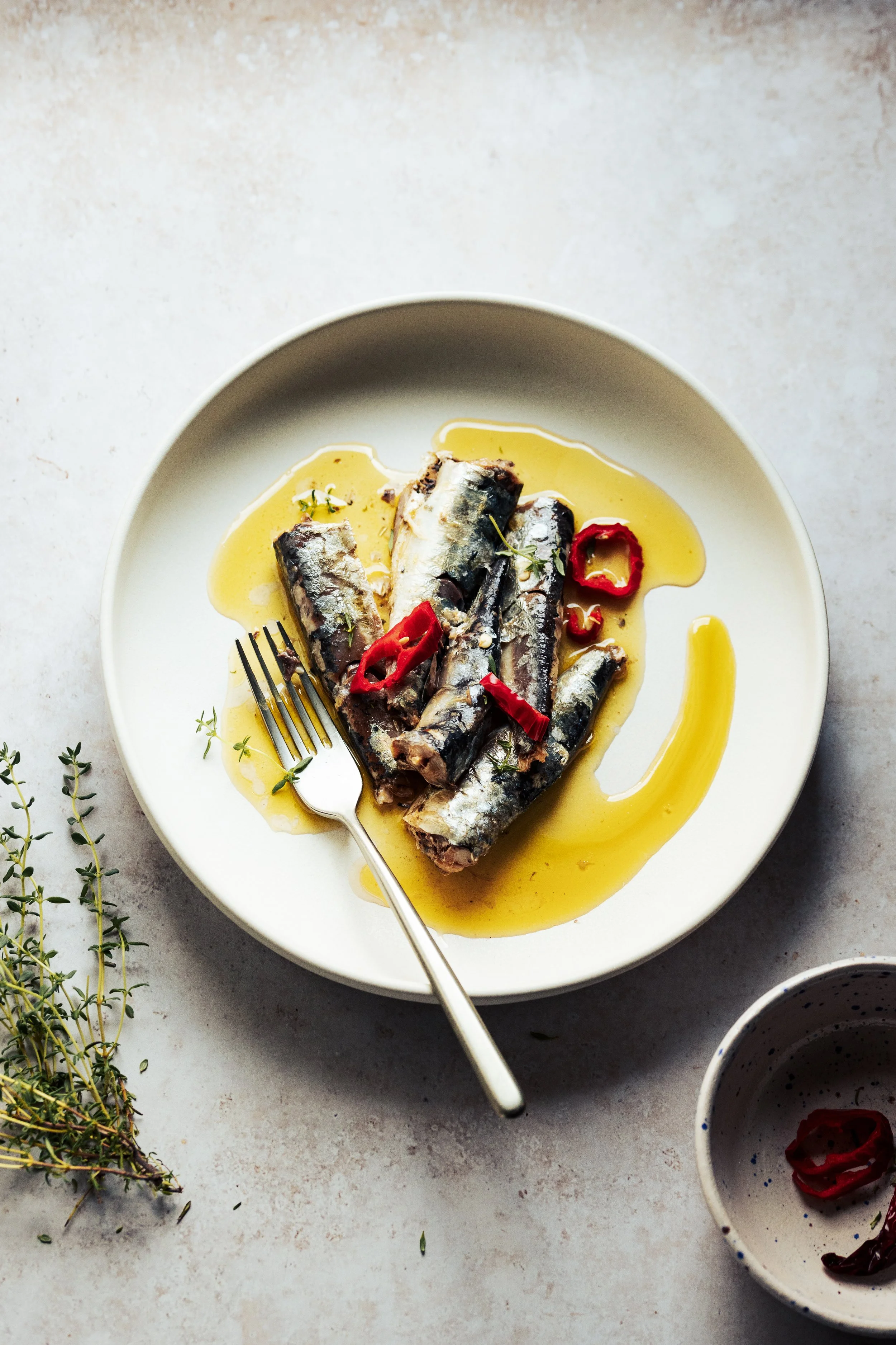 Bowl of cooked sardines in lemon sauce garnished with red chili slices and herbs, with two cups of red wine and sprigs of herbs on a white surface.