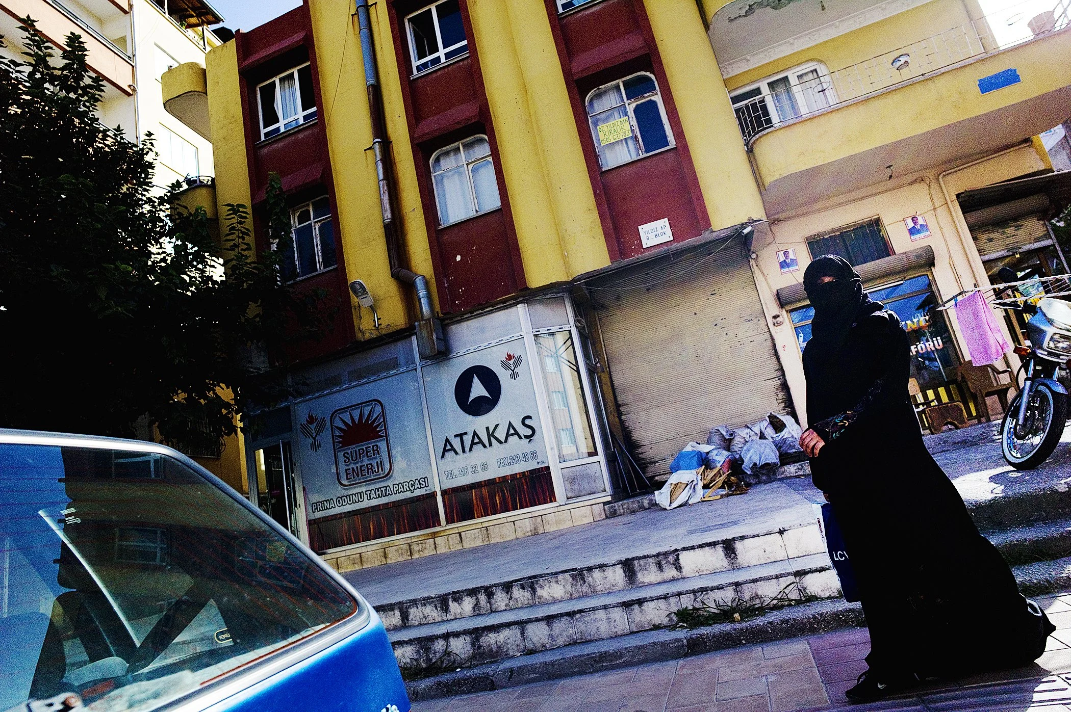 A person wearing a black hooded jacket and black face covering walking past a yellow and red apartment building with shops on the ground floor. The building has some signs and posters, including a large truck with Turkish text parked in front. A tree partially blocks the left side of the scene, and part of a blue vehicle is visible in the foreground.