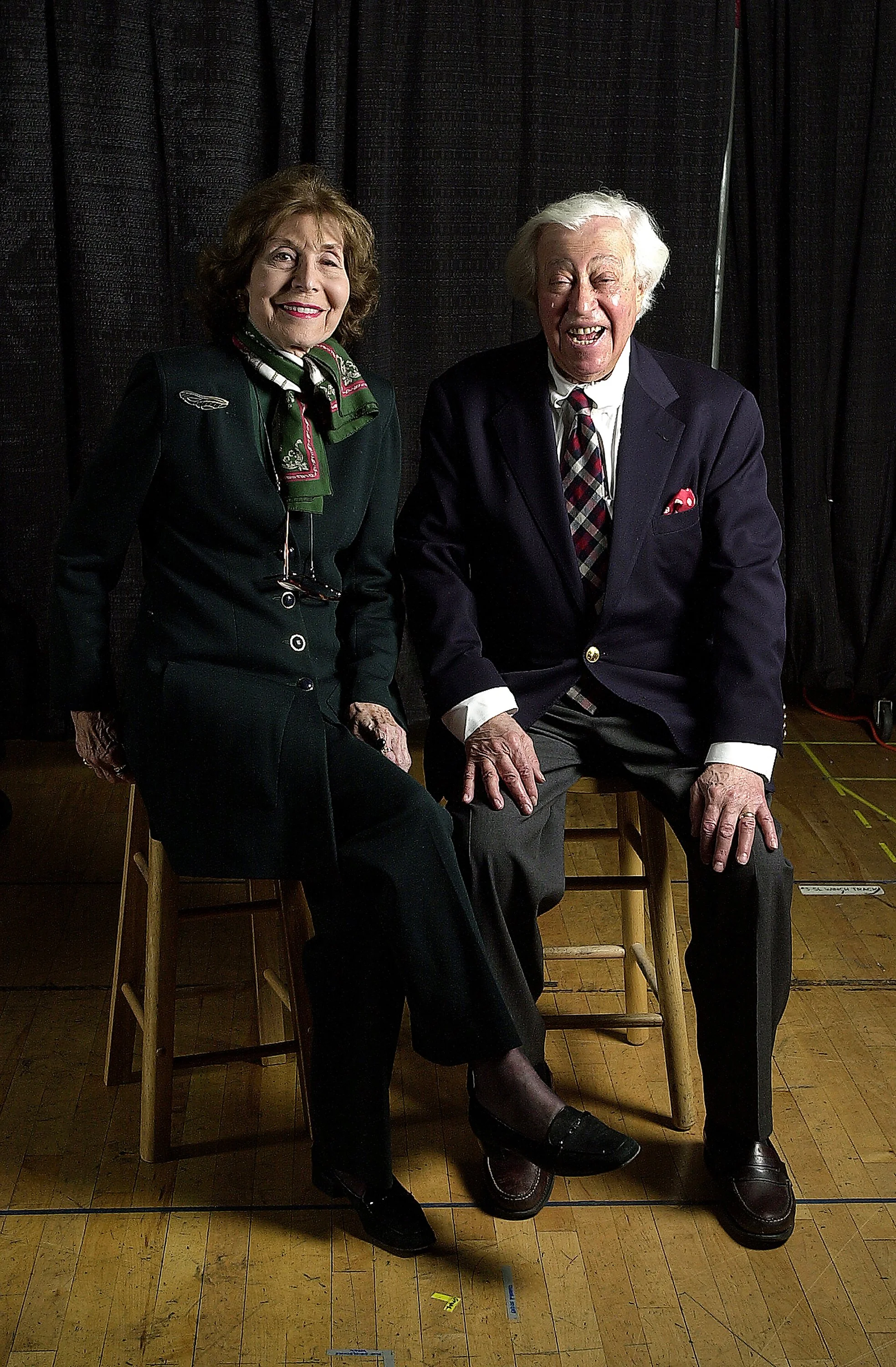 A woman and an elderly man smiling and sitting on chairs in front of a black curtain. They are dressed in formal clothing with the woman wearing a dark suit, green scarf, and glasses hanging from her neck, and the man wearing a navy blazer with a red