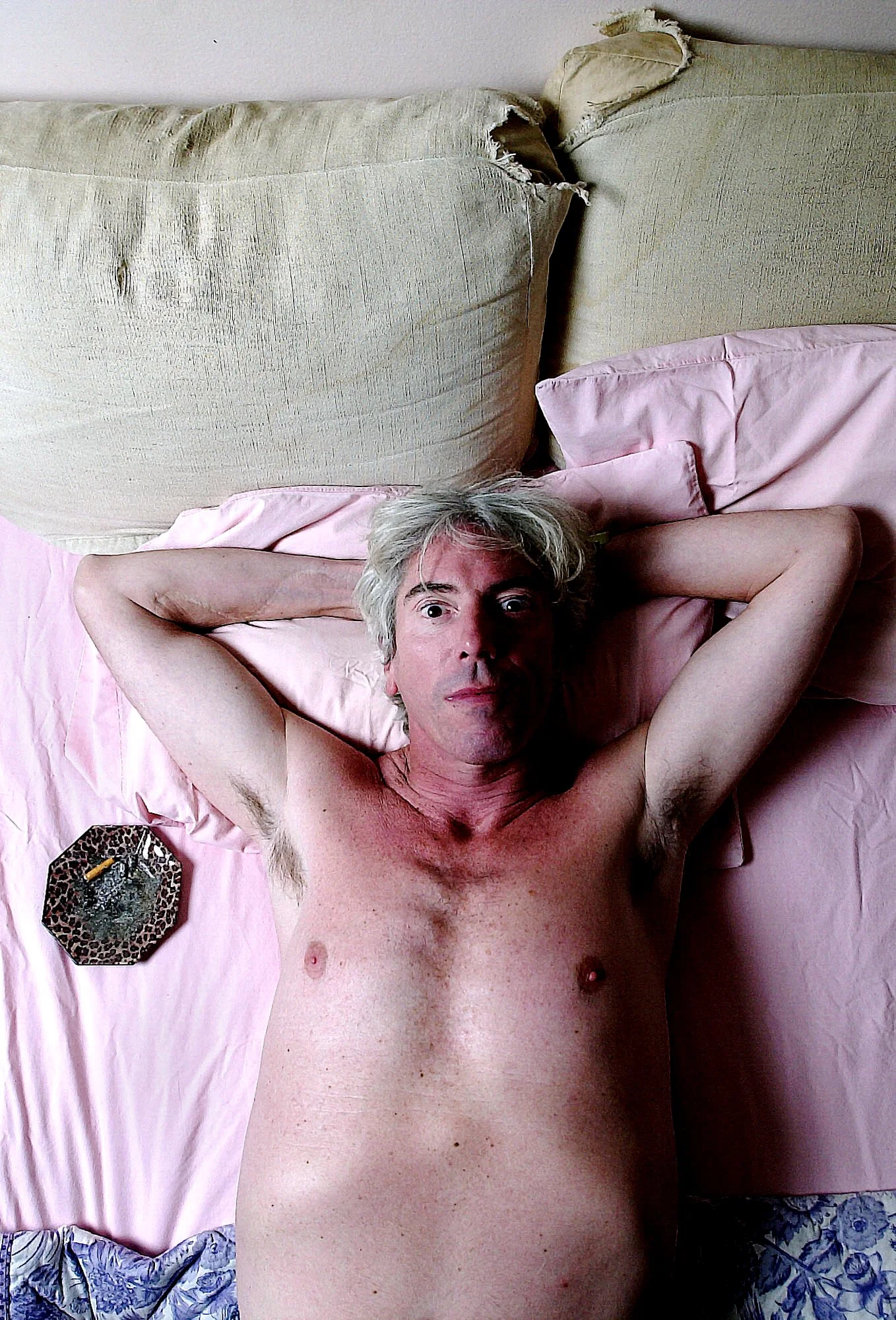 A topless middle-aged man with gray hair laying on a bed with pink sheets, hands behind head, looking at the camera. There are two pillows behind his head, a leopard print ashtray with a lit cigarette on the left side of the bed, and a blue patterned
