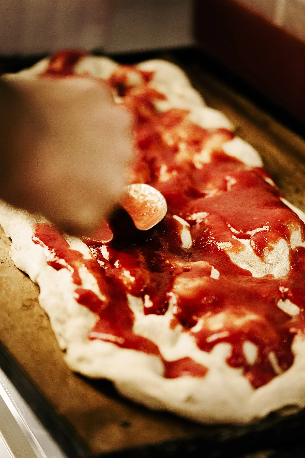 A person spreading tomato sauce on pizza dough with a spoon.