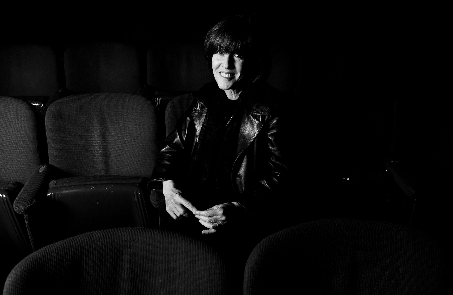 A woman with short dark hair sitting in a dark auditorium with empty seats around her, wearing a leather jacket and smiling at the camera.