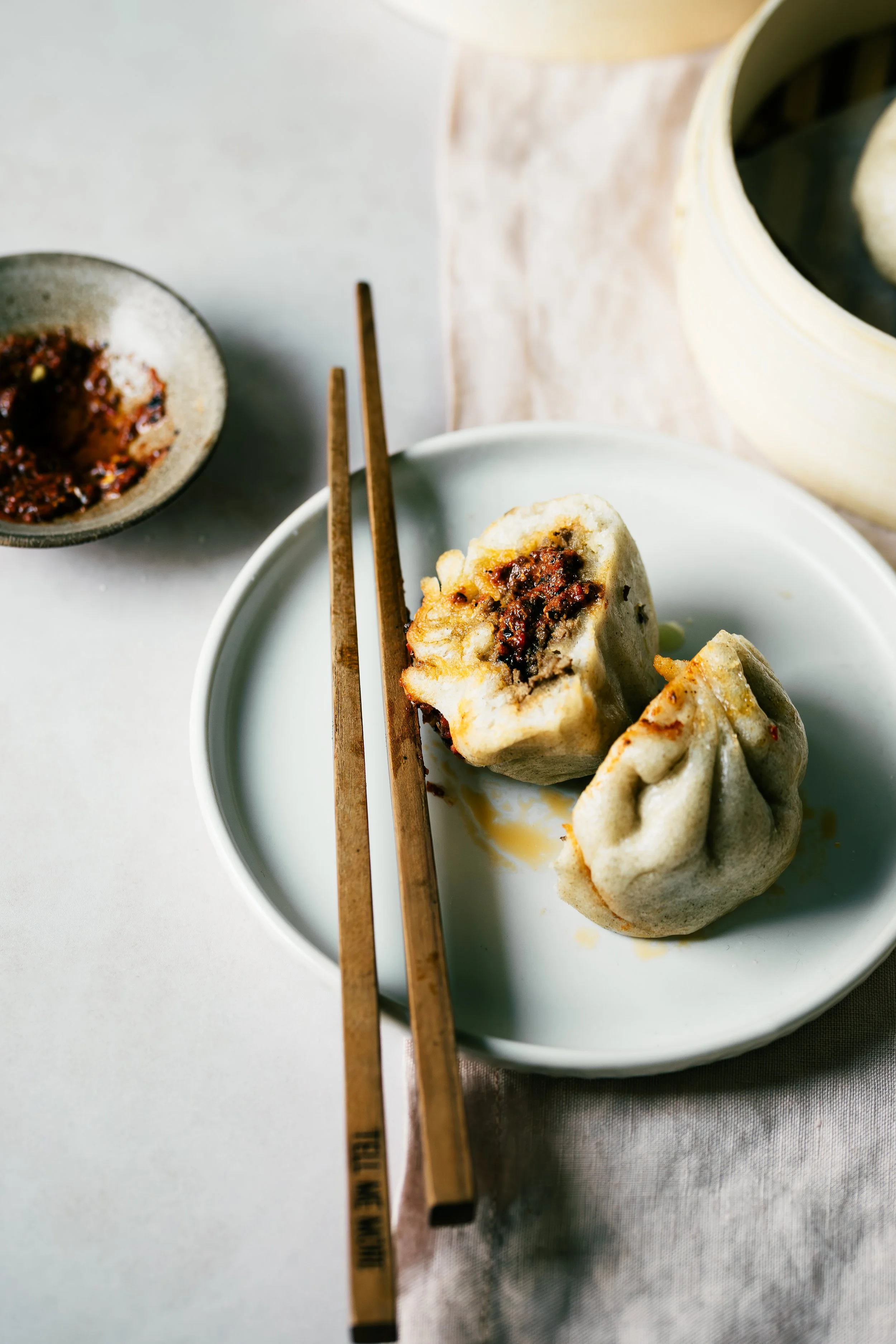 Close-up of two steamed dumplings on a white plate with dipping sauce in a small bowl and chopsticks resting on the plate.