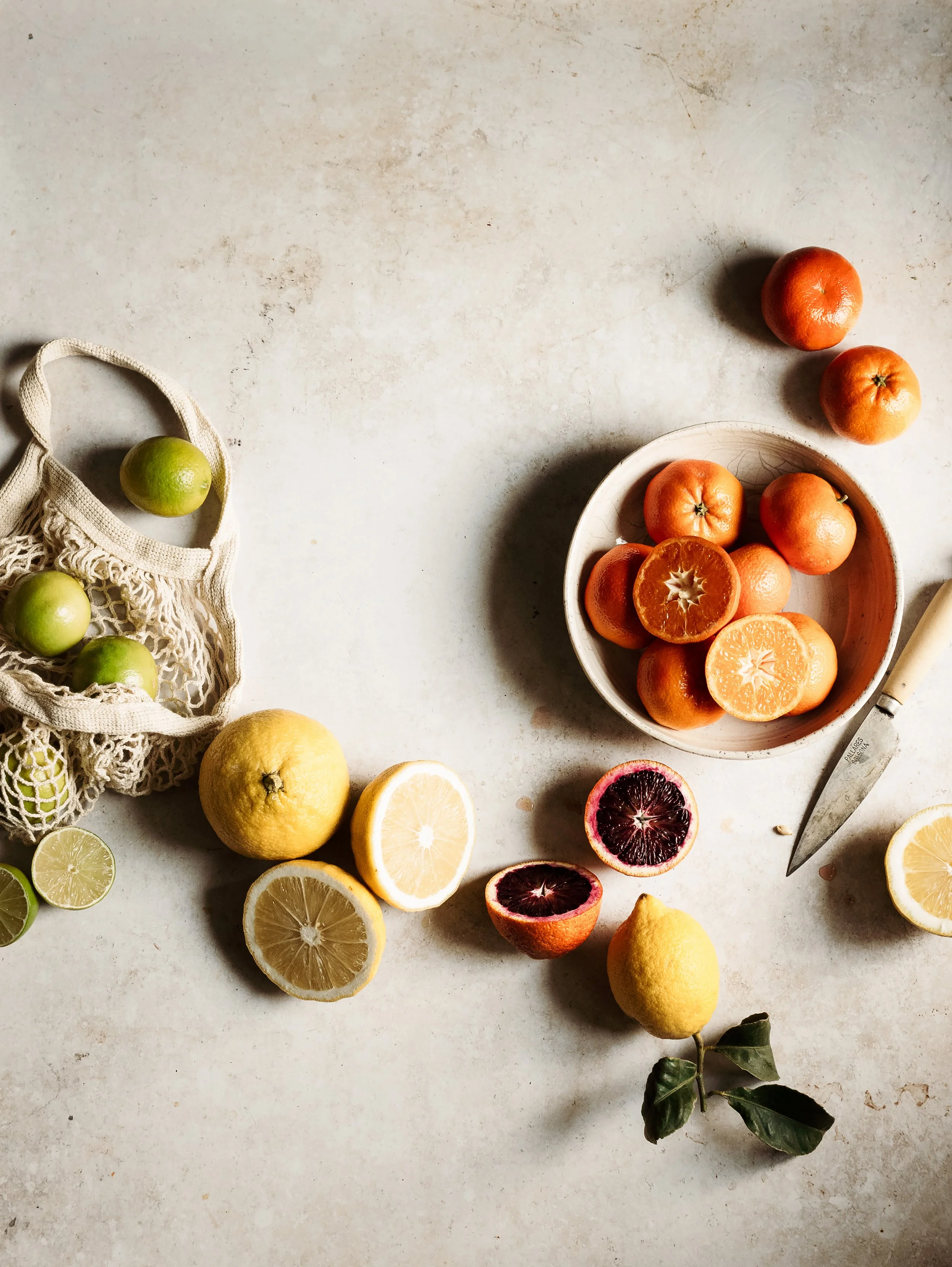 Assorted citrus fruits on a light-colored surface, including lemons, limes, oranges, and a blood orange, with some cut open showing their interiors. A grocery bag with green limes and a knife are also visible.
