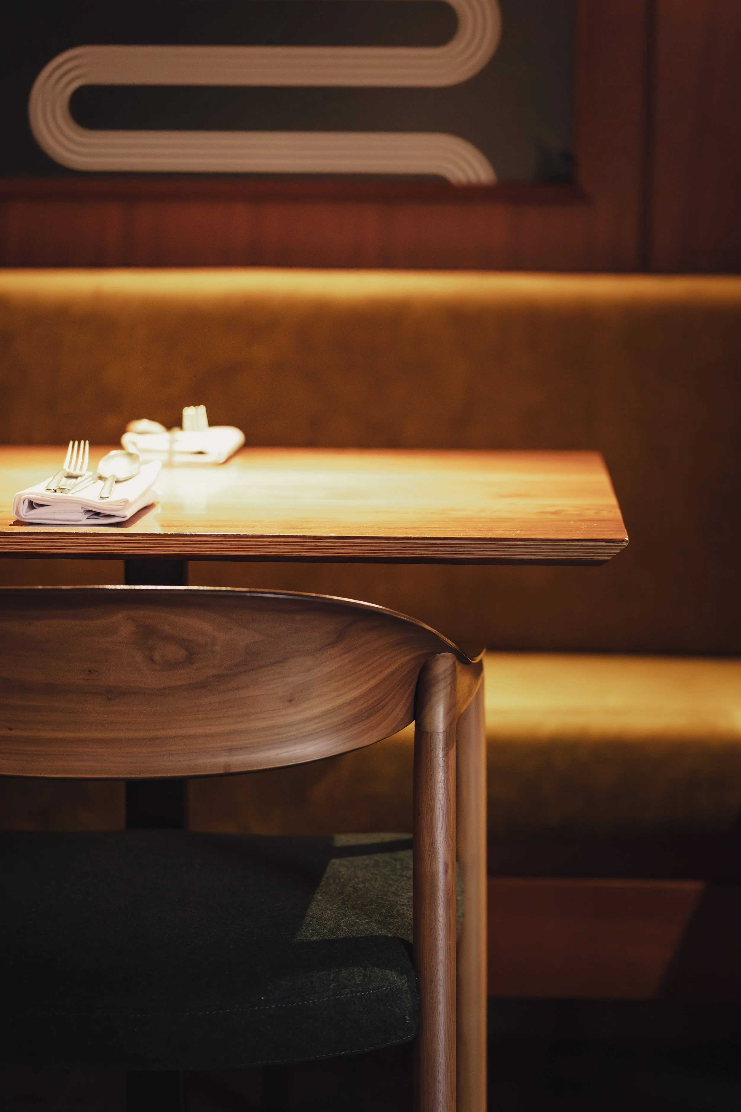An empty restaurant table set with silverware and napkins, with a wooden chair in the foreground and a booth in the background.