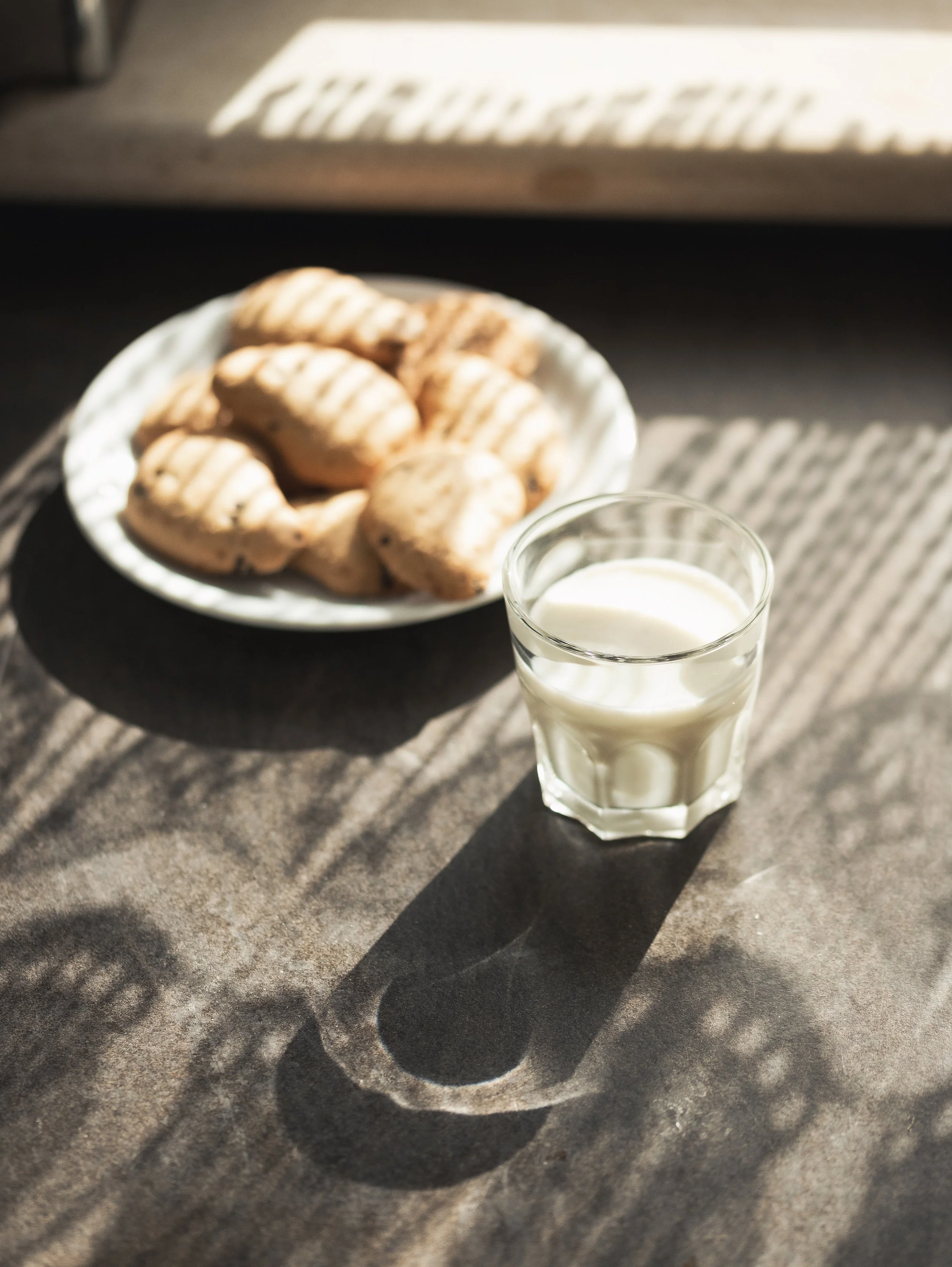 A glass of milk and a plate of cookies on a dark surface with sunlight creating shadows.