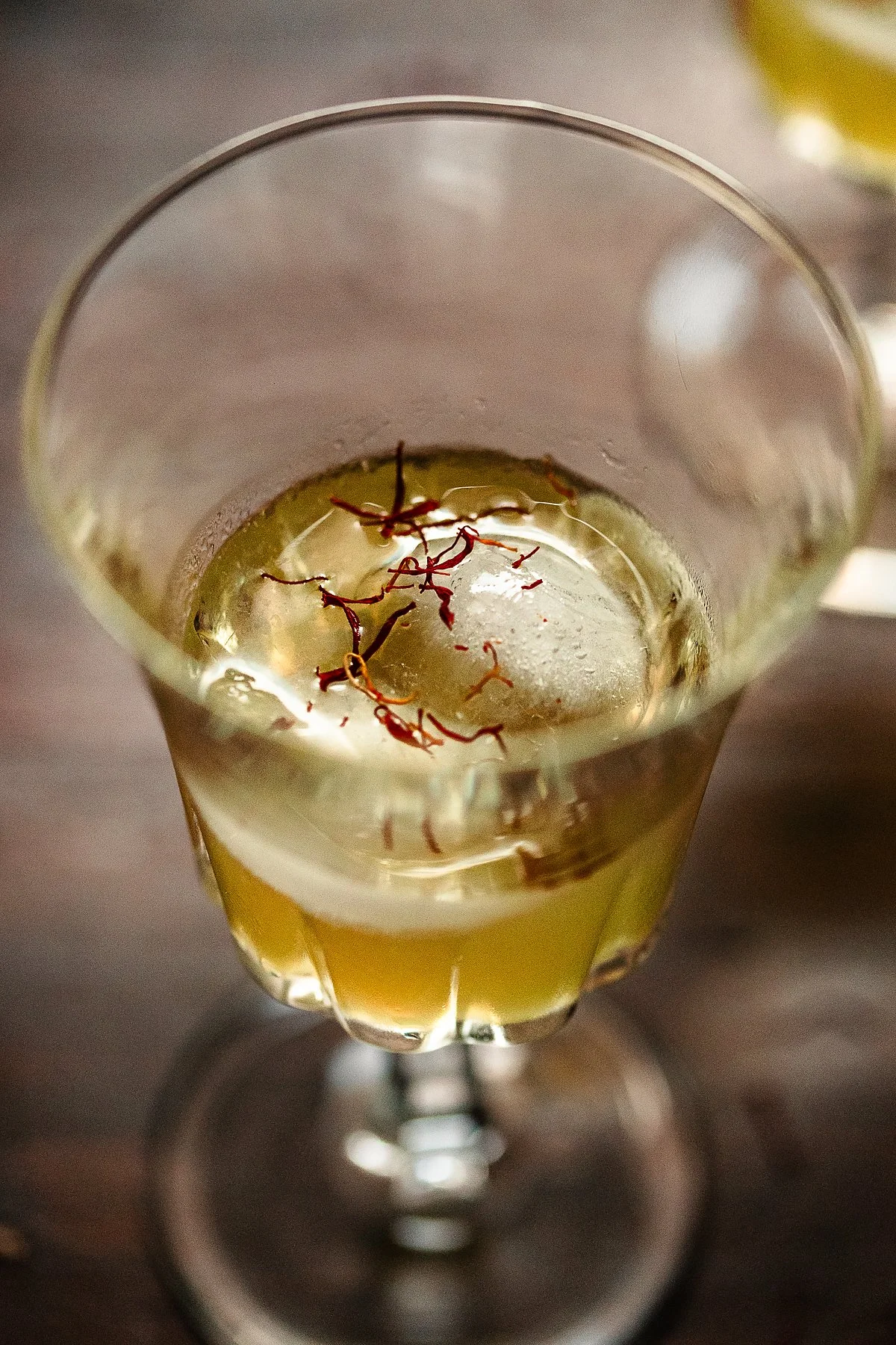A close-up view of a glass of white wine garnished with red saffron threads, placed on a wooden surface.