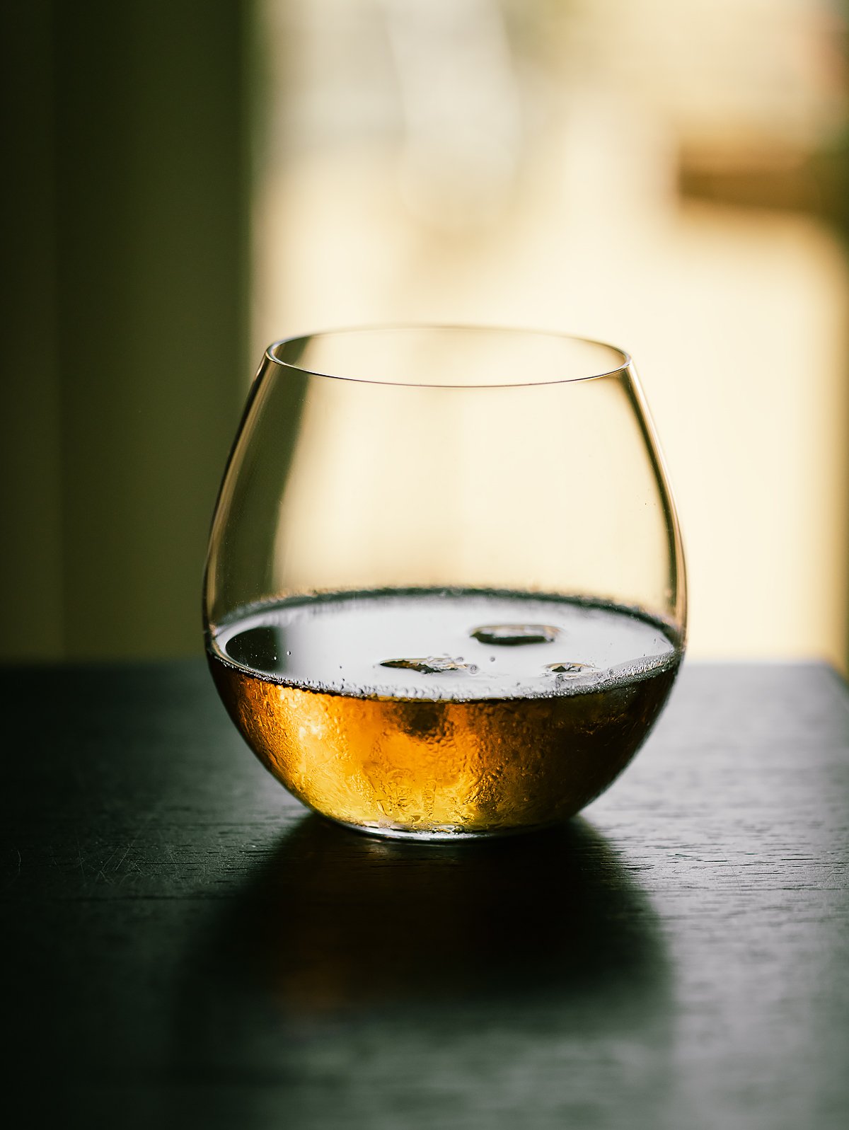 A glass of amber-colored beverage with ice cubes on a dark surface, with a blurred light background.