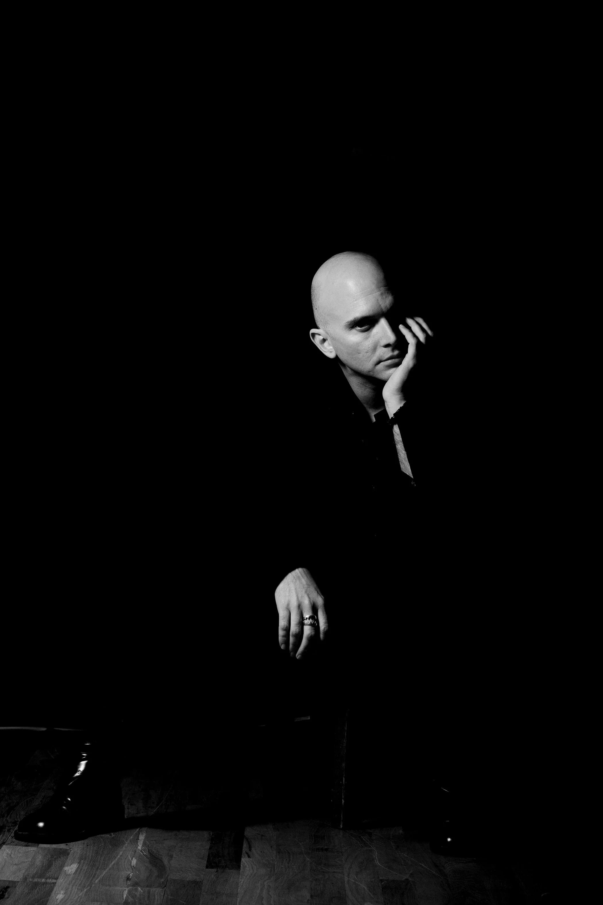 Black and white photo of a bald man with intense expression, resting his face on his hand, sitting in a dark room with a black background, wearing rings on his fingers and dark clothing.