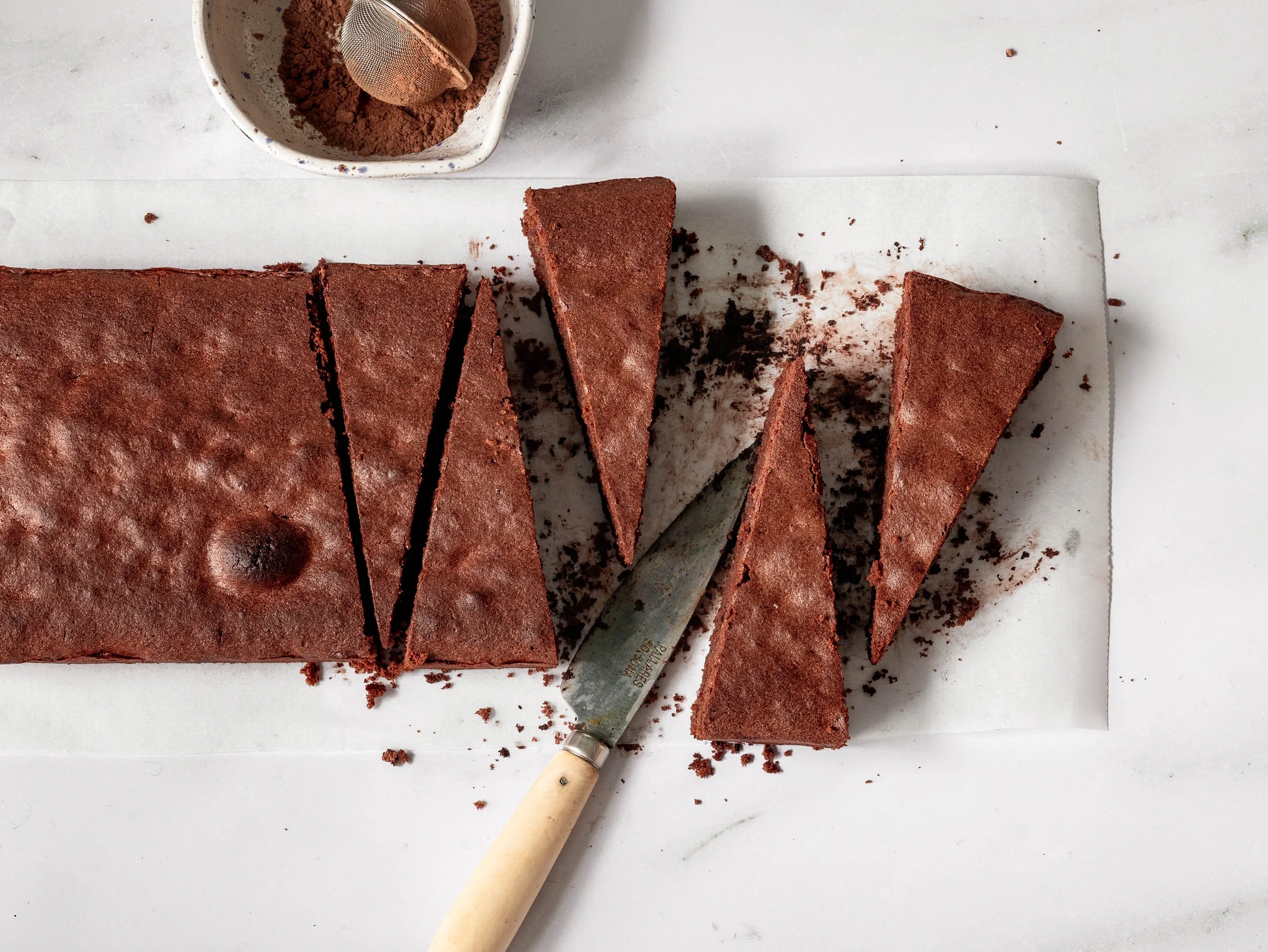 A sliced chocolate cake on white parchment paper with crumbs scattered around, and a spoon in the cake. A bowl containing cocoa powder or flour with a sifter is in the background.