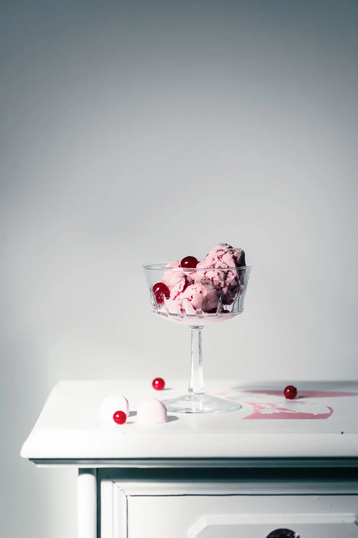 A glass bowl of cherry-flavored ice cream with cherries on top, placed on a white surface with a few cherries and a marshmallow around it, against a plain gray background.