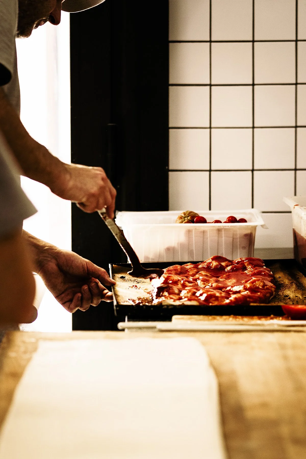 Person cutting a pizza topped with tomato sauce on a wooden countertop in a kitchen.