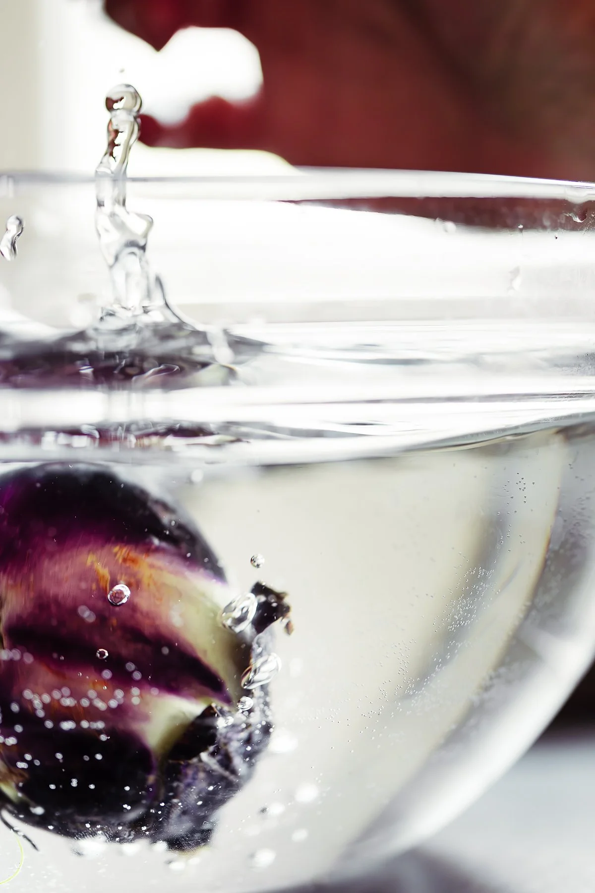 Close-up of a dark purple berry being dropped into a glass of clear water, creating bubbles and water splashes.