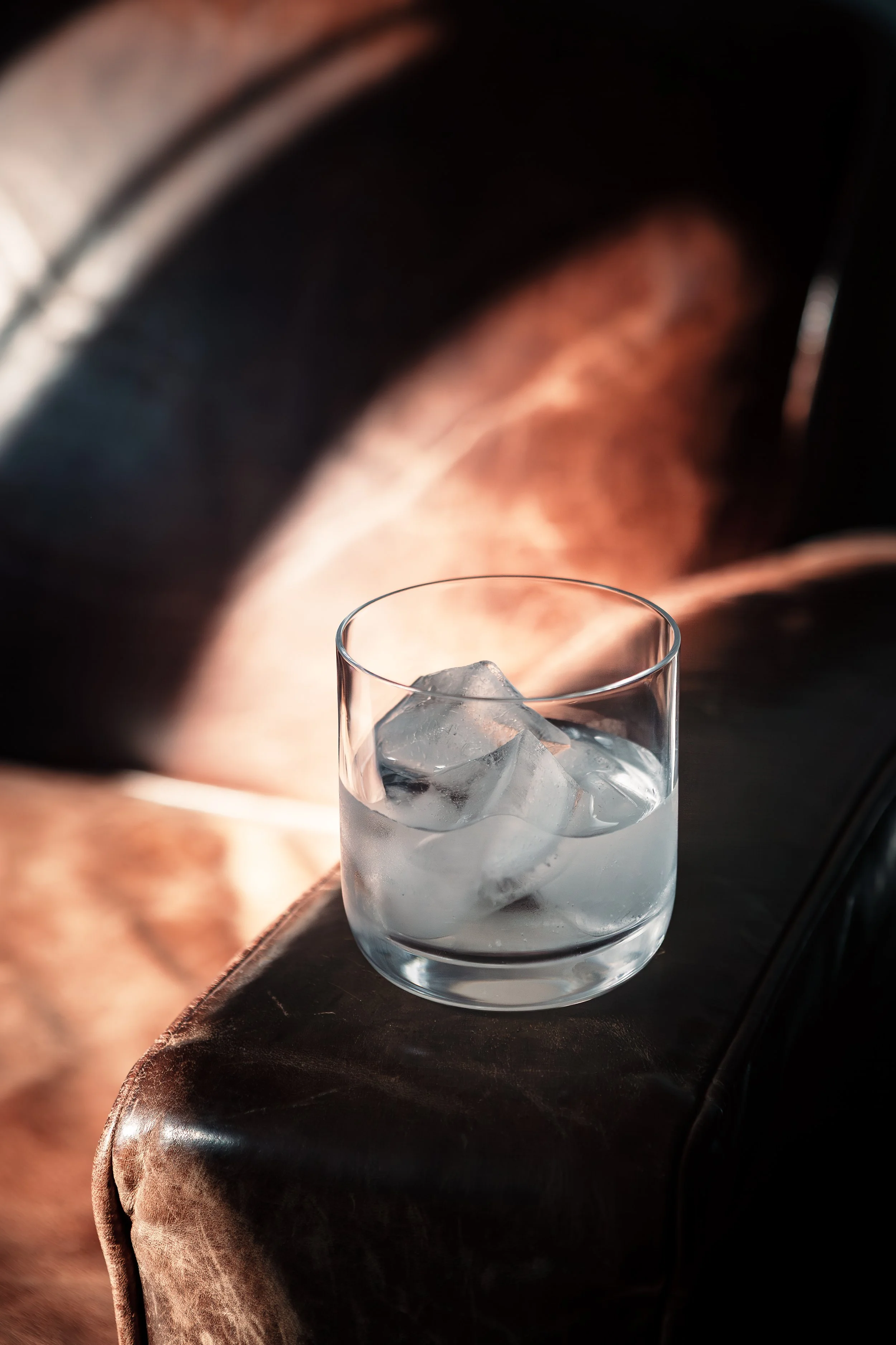 A glass of water with ice cubes on a dark surface, with a blurred background featuring a black leather armrest and warm lighting.
