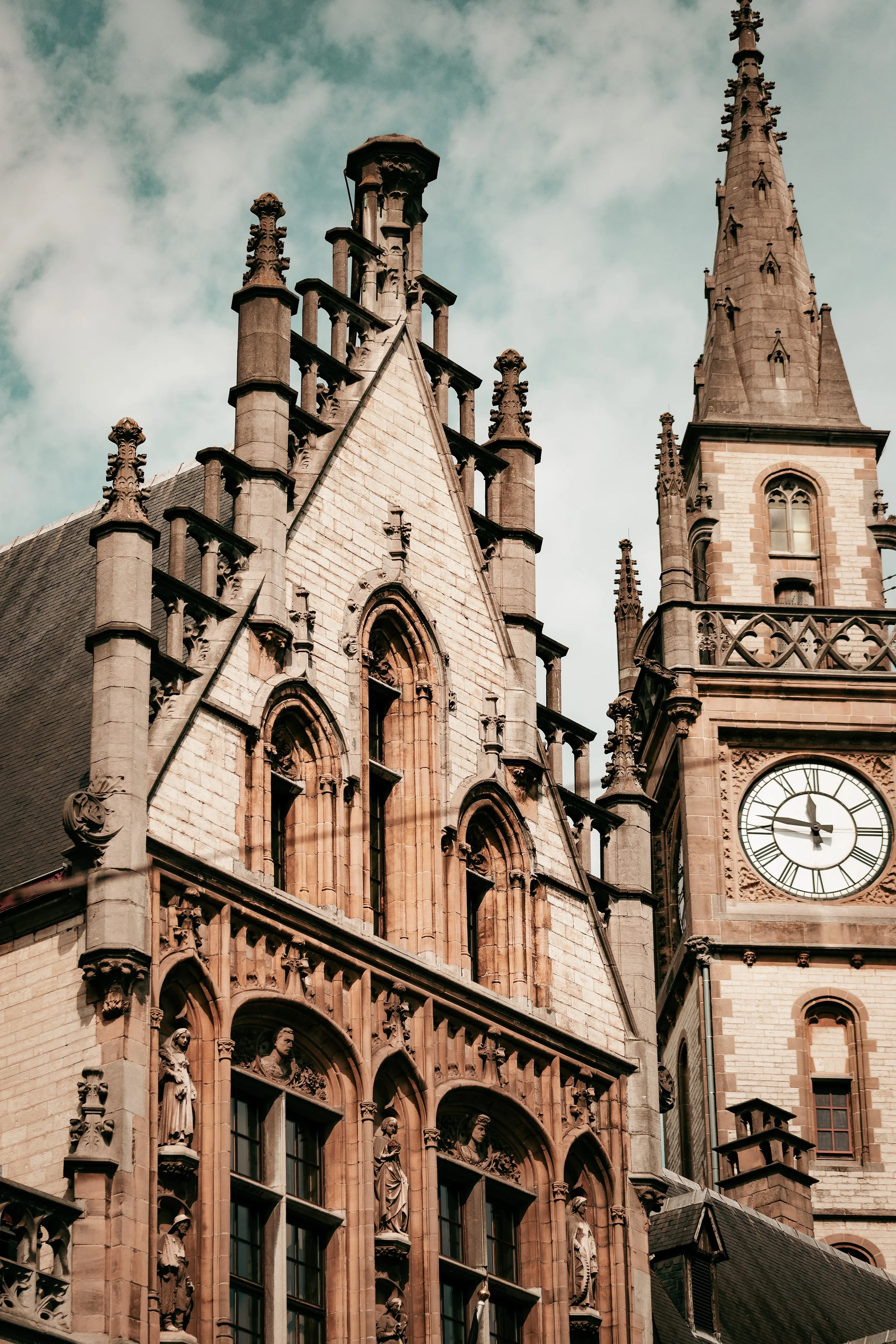 A historic Gothic-style church or cathedral with intricate stone carvings, arched windows, and a clock tower, set against a partly cloudy sky.