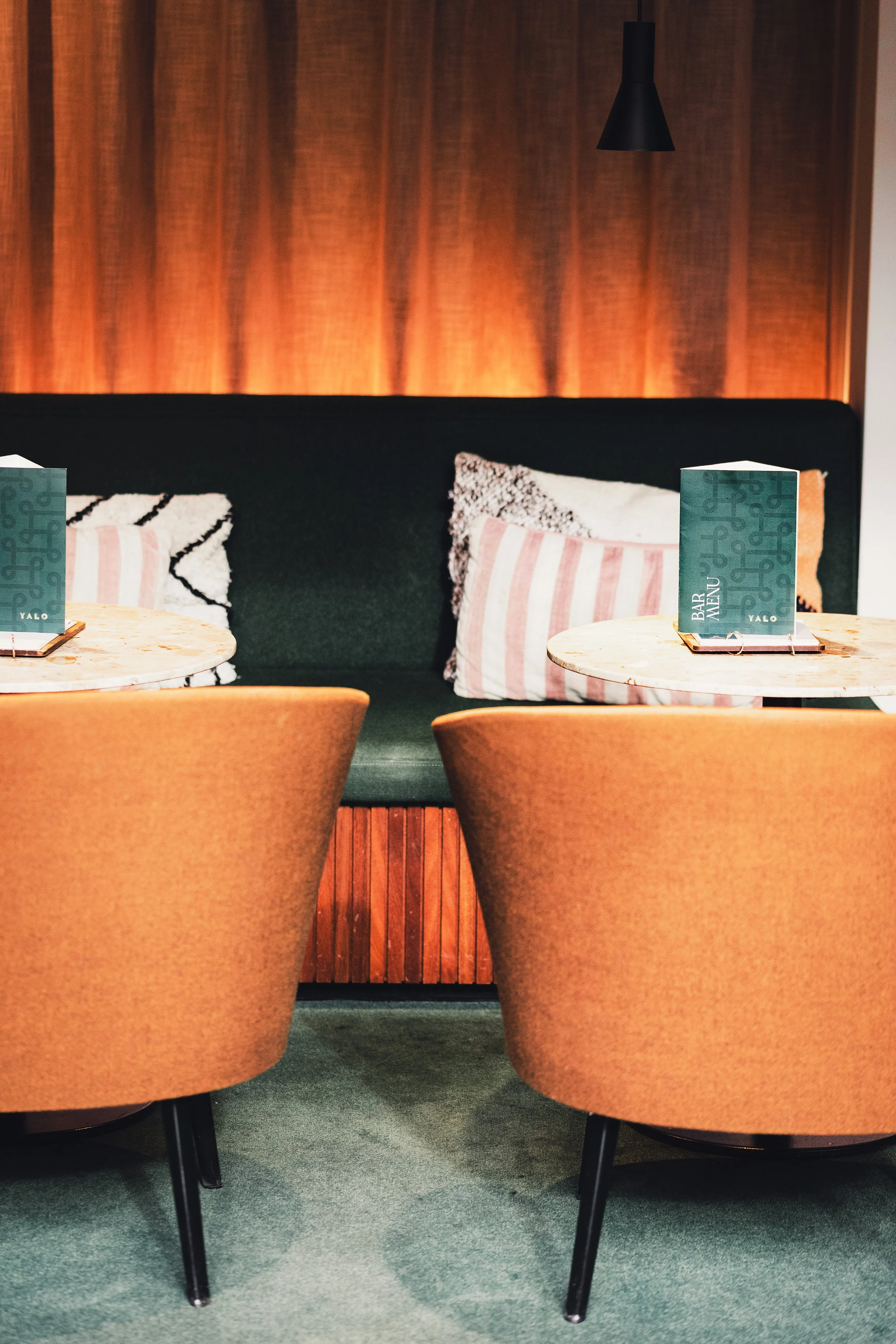 Interior view of a cozy restaurant with two round tables, each with a menu and chairs, a cushioned bench with pillows in the background, and a modern black pendant light hanging from the ceiling.