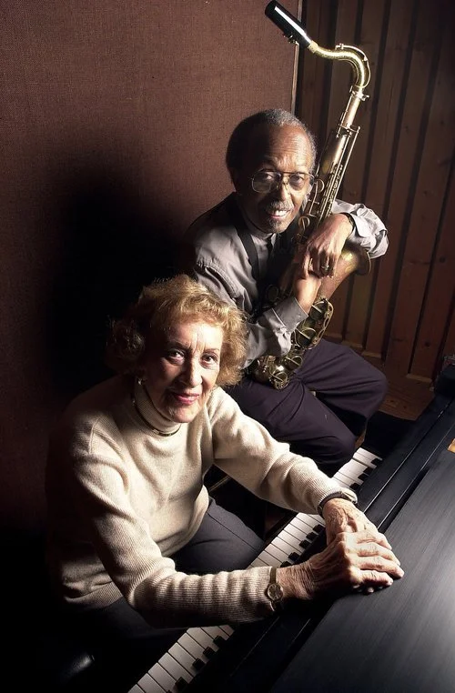 An elderly woman sitting at a piano with her hands on the keys, and a man sitting behind her holding a saxophone, both smiling at the camera in a music studio.