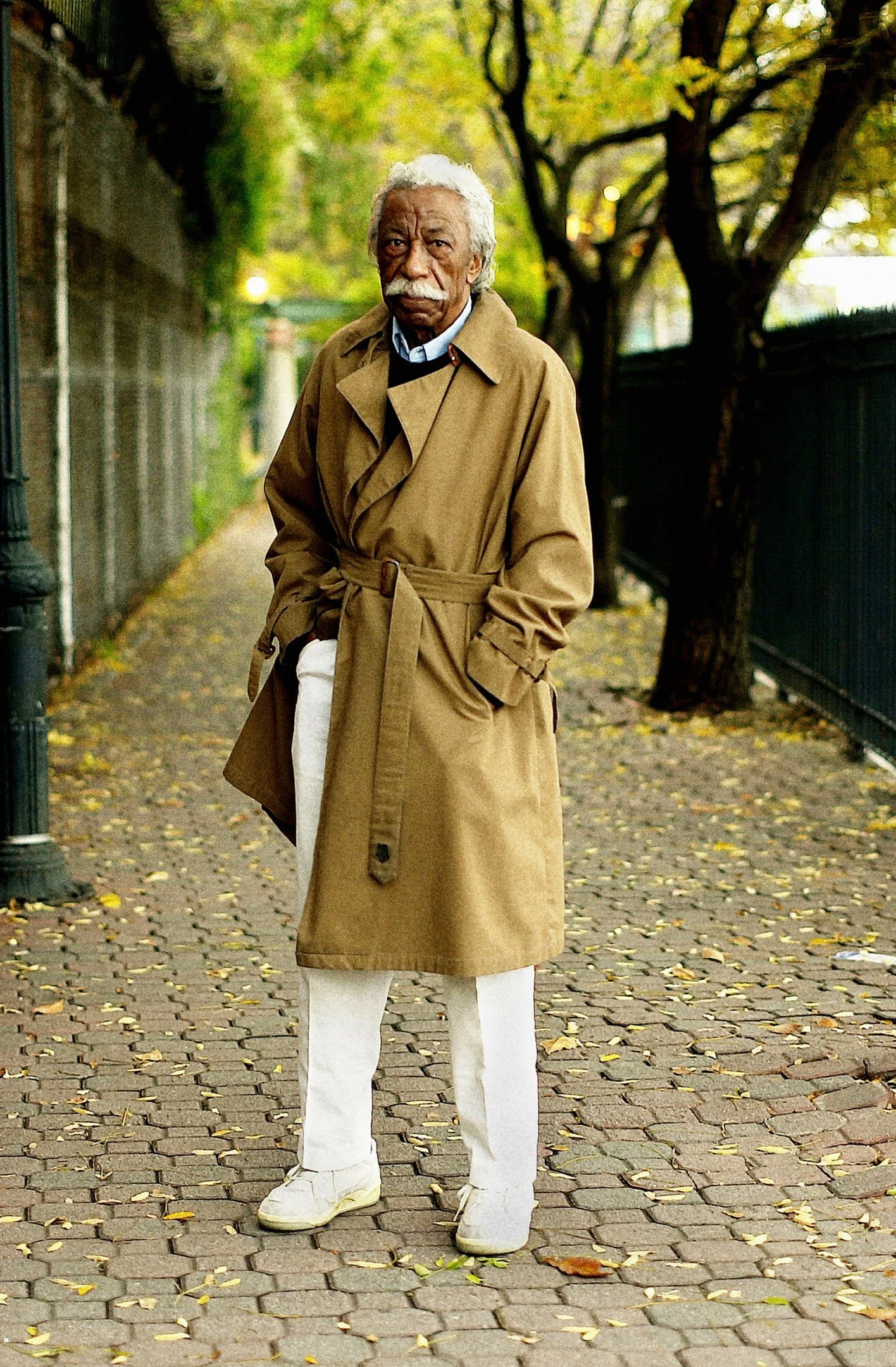 An elderly man with white hair and a mustache standing on a leaf-covered cobblestone sidewalk, wearing a long beige trench coat and white pants with sneakers, under trees with autumn leaves.