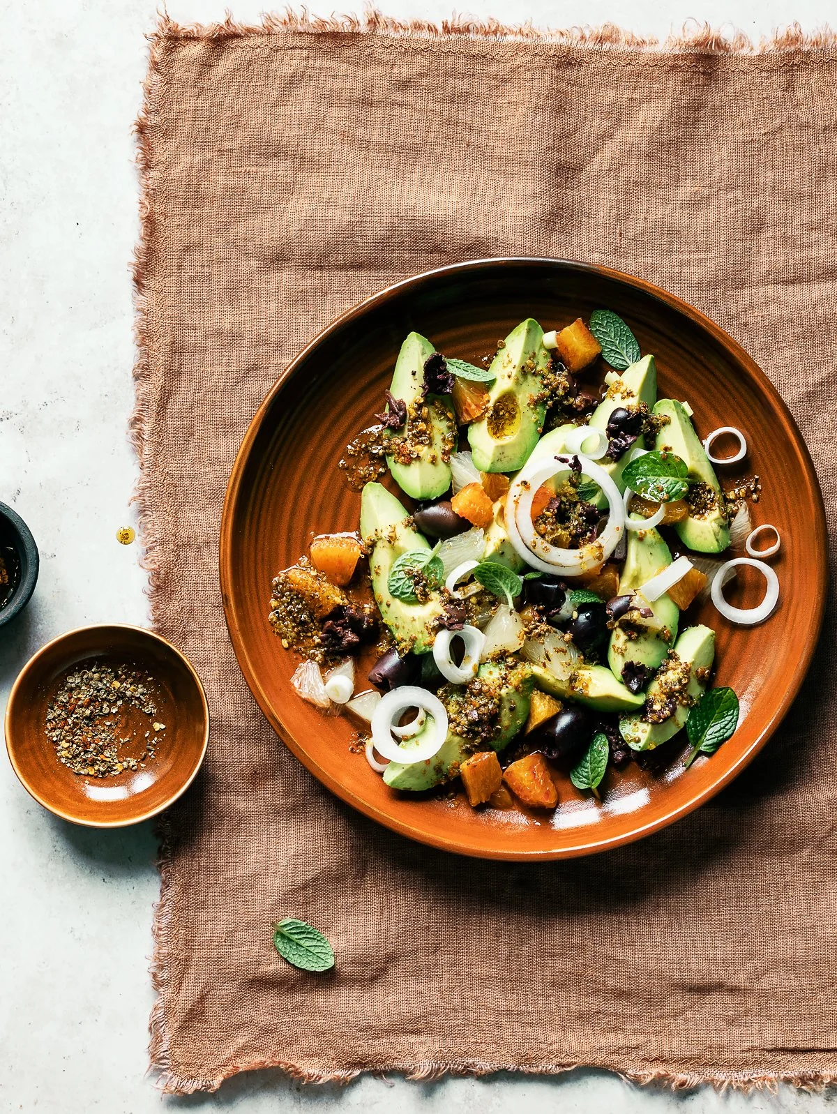 A salad with avocado, onion, herbs, black olives, and roasted vegetables on a brown plate.