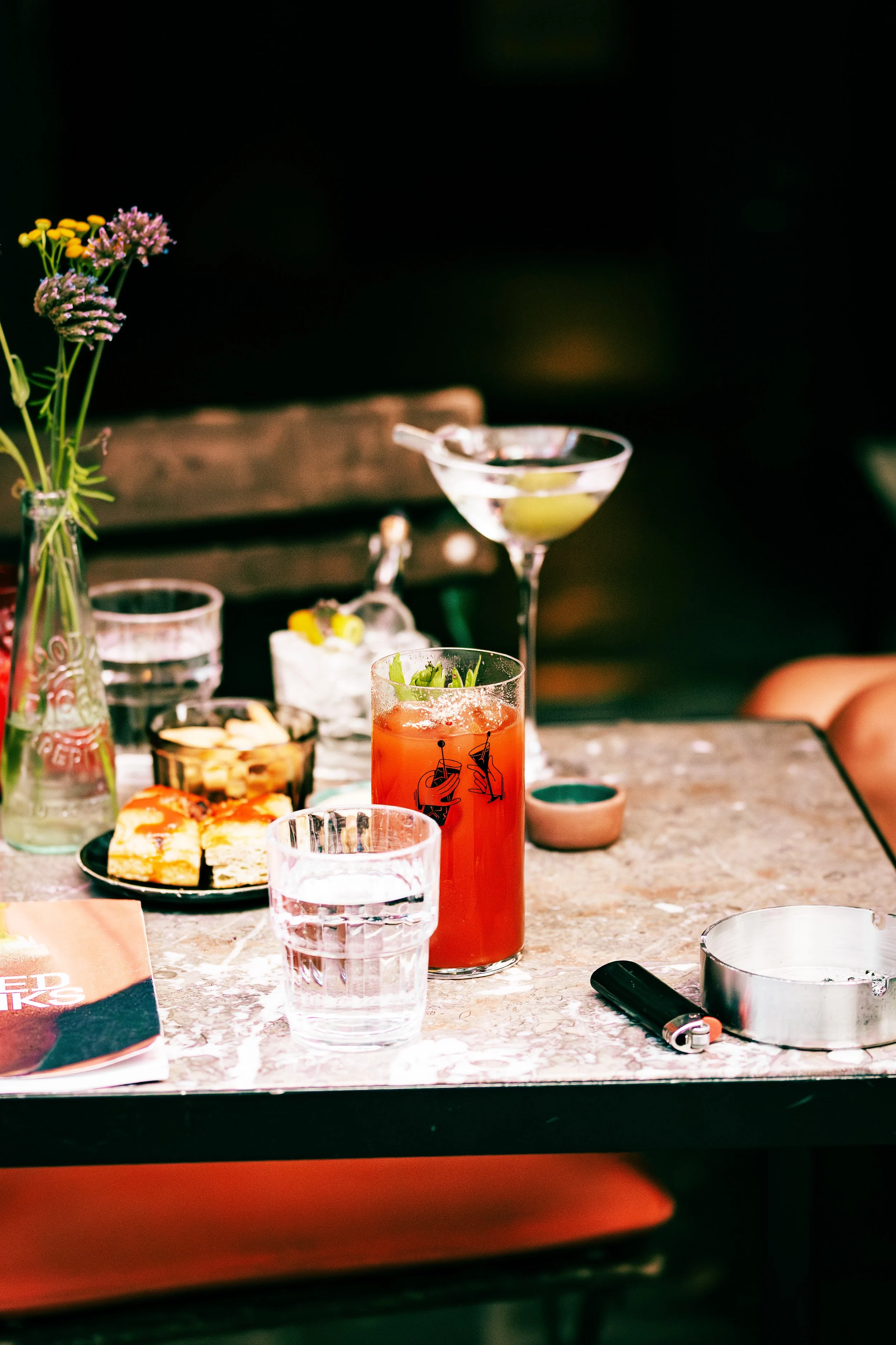 A table with drinks including a glass of water, a bloody mary, a cocktail with a slice of lemon, and a martini, with a plate of pizza, a book, a small plant, and a lighter, set against a dark background.
