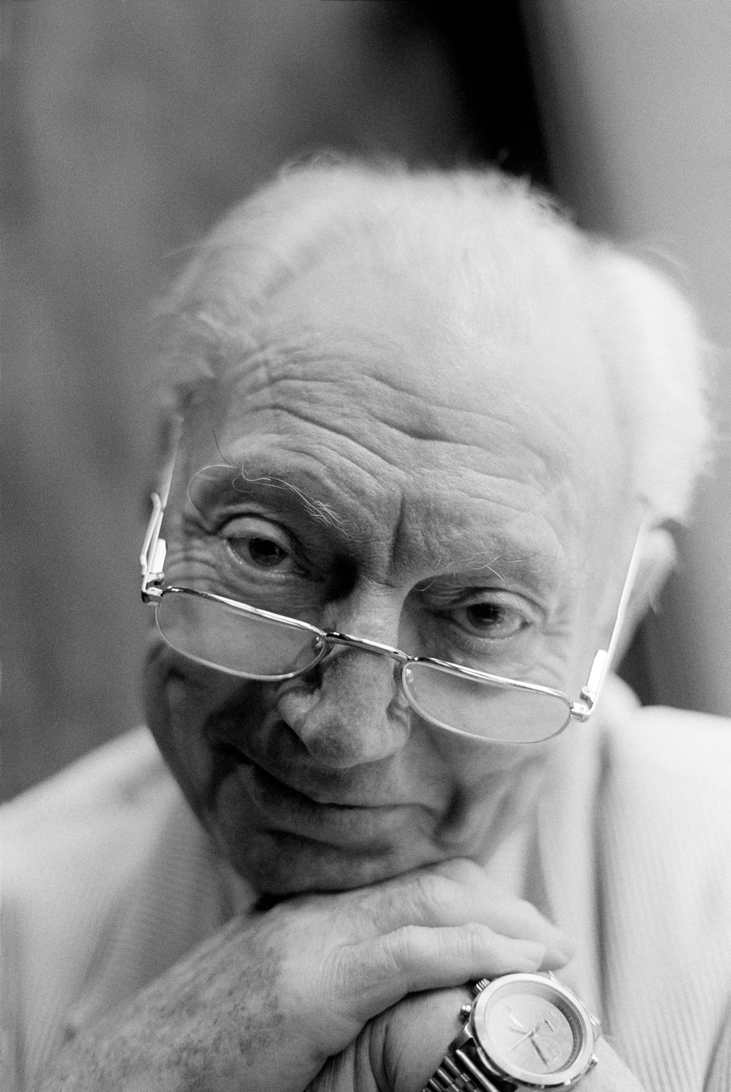 Close-up of an elderly man looking towards the camera, wearing glasses, a watch, and a hoodie, with his hand partially covering his face.