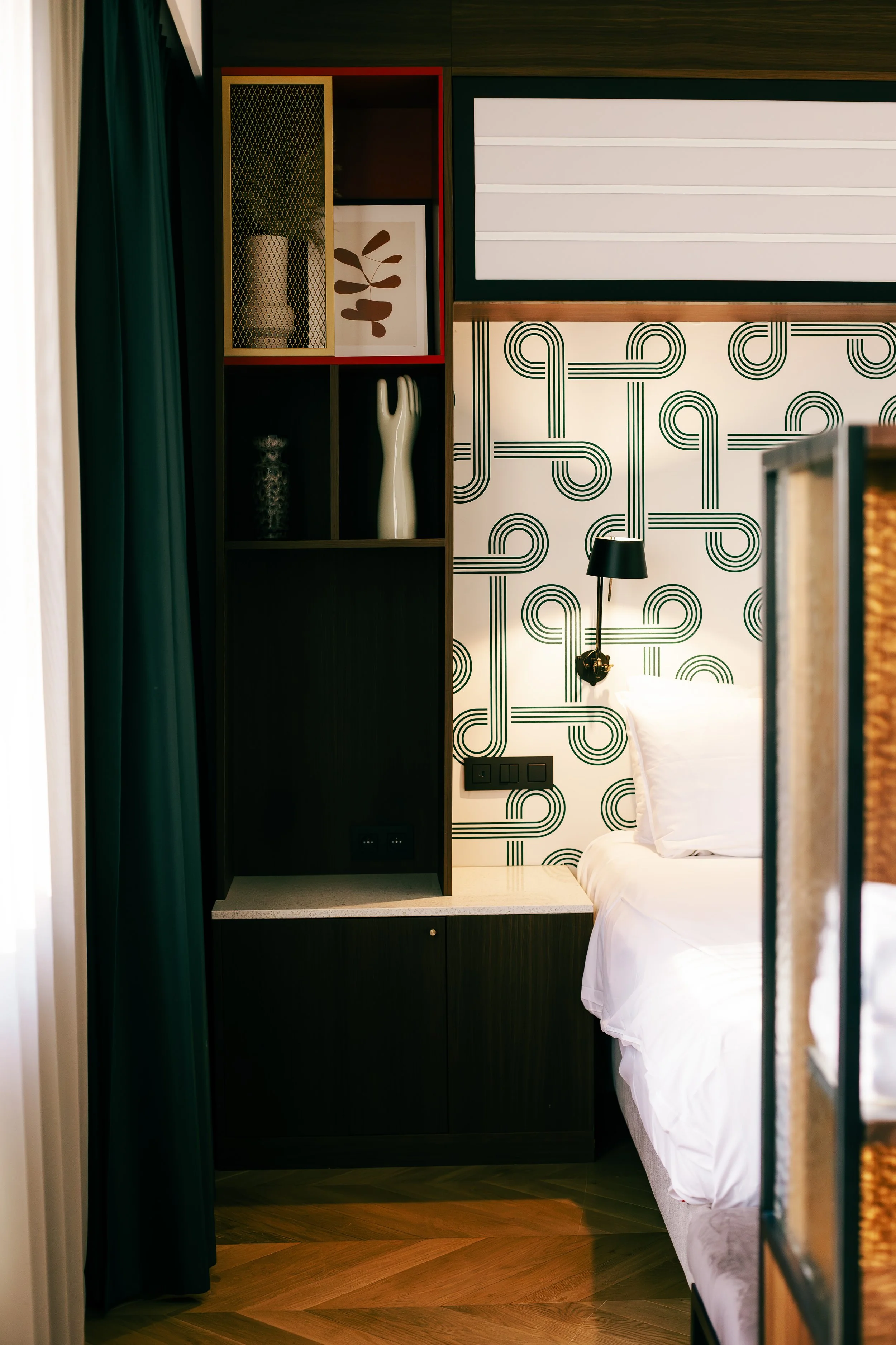 Close-up view of a modern hotel room with a bed, black wall-mounted reading lamp, textured wallpaper, and a black shelving unit with decorative items.