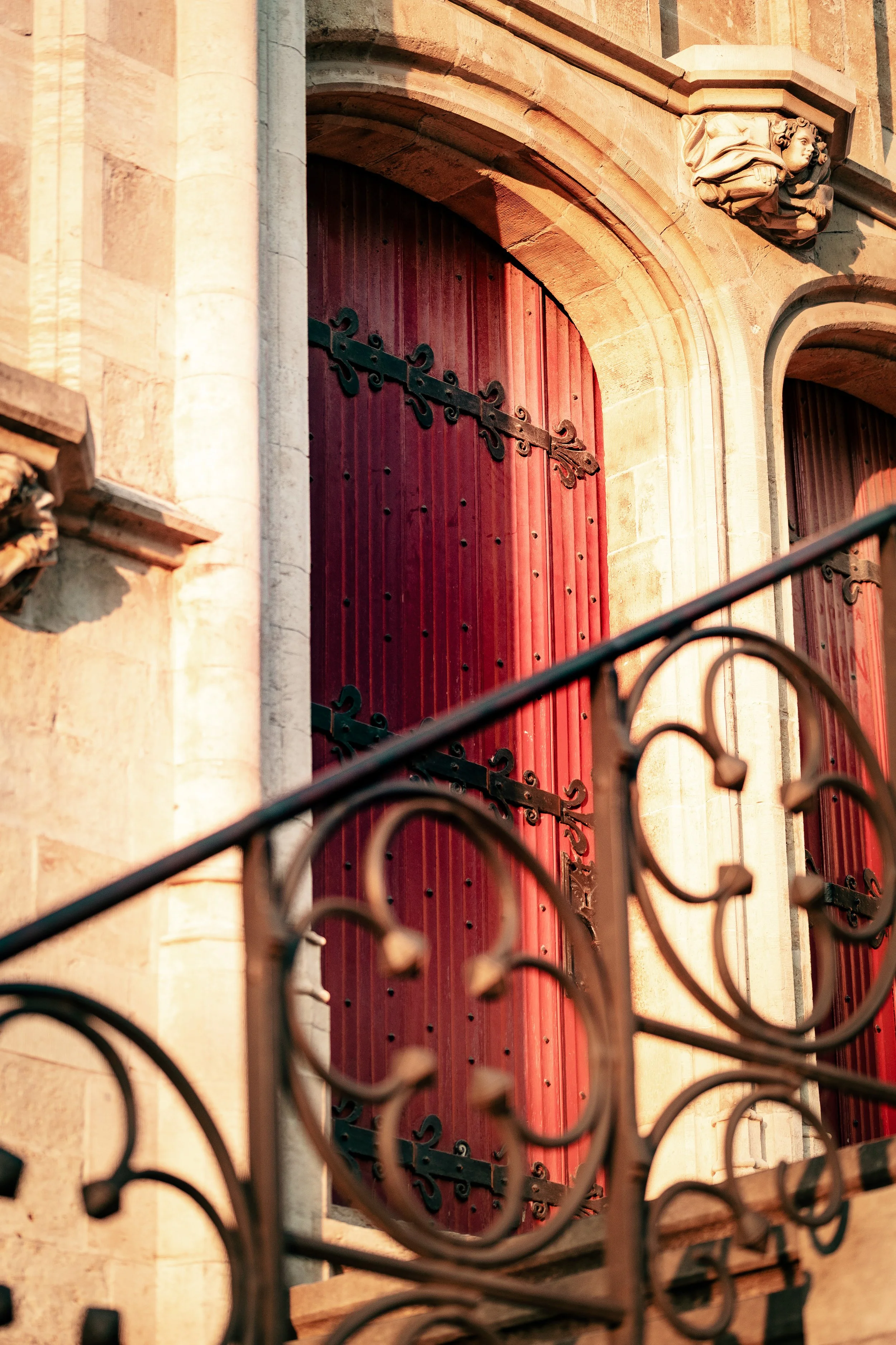 Close-up of a historic building with red wooden shutters, decorative iron hinges, and ornate stone architectural details. Sunlight casts a warm glow.