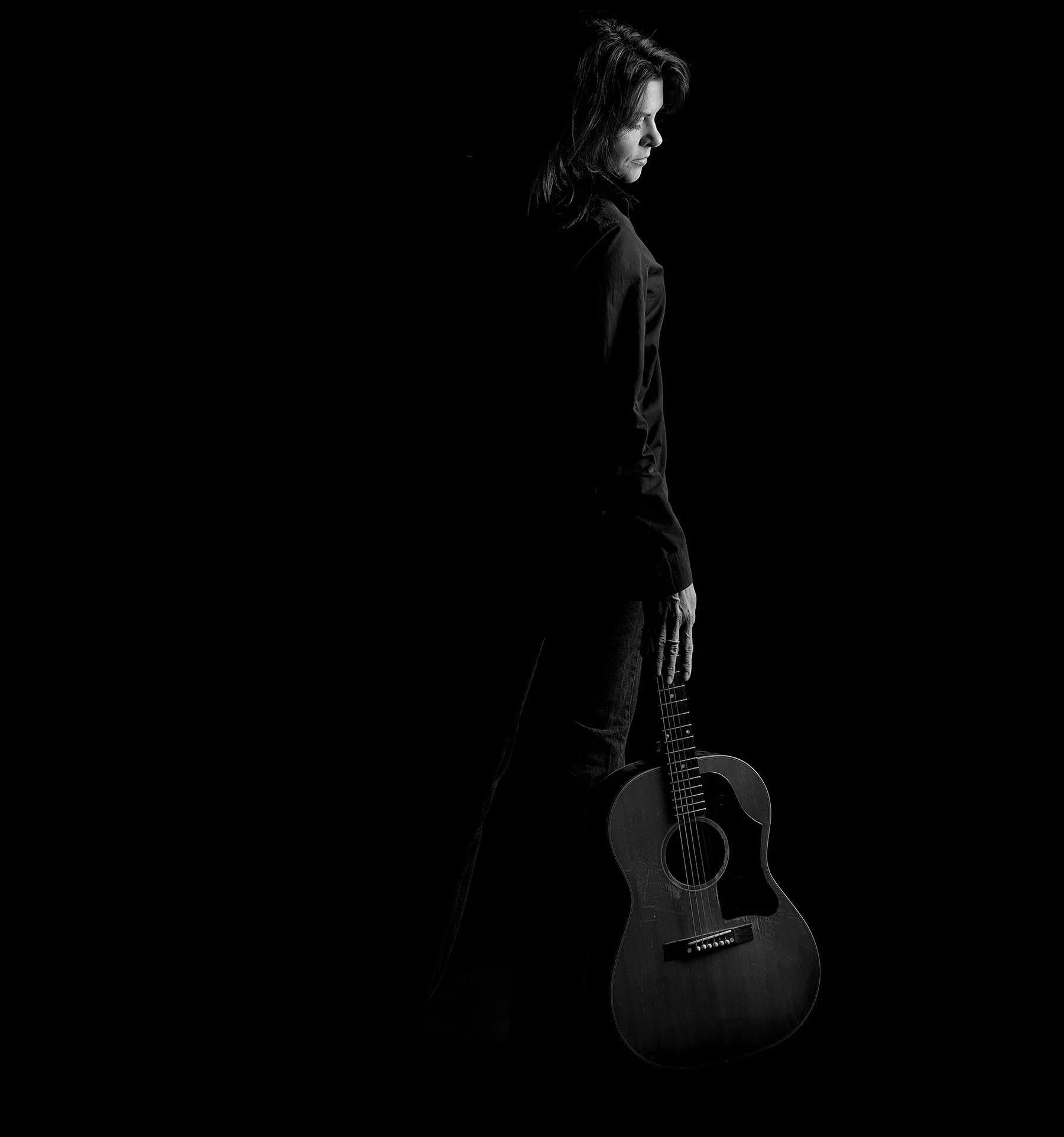 Black and white photo of a woman with long hair, standing in profile, holding an acoustic guitar in her right hand, with a dark background.