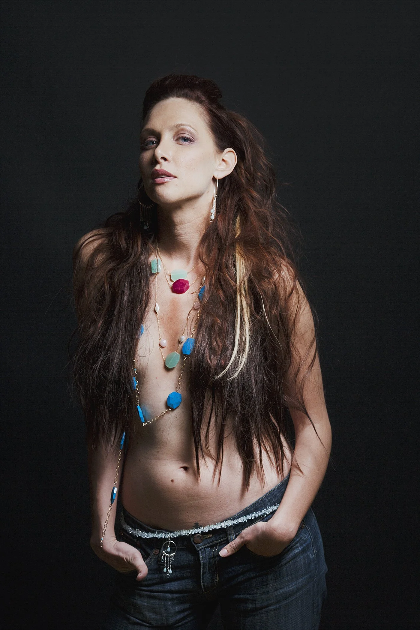 A woman with long, wavy red hair poses shirtless against a dark background, wearing multiple colorful necklaces and earrings, with her left hand in her pocket.