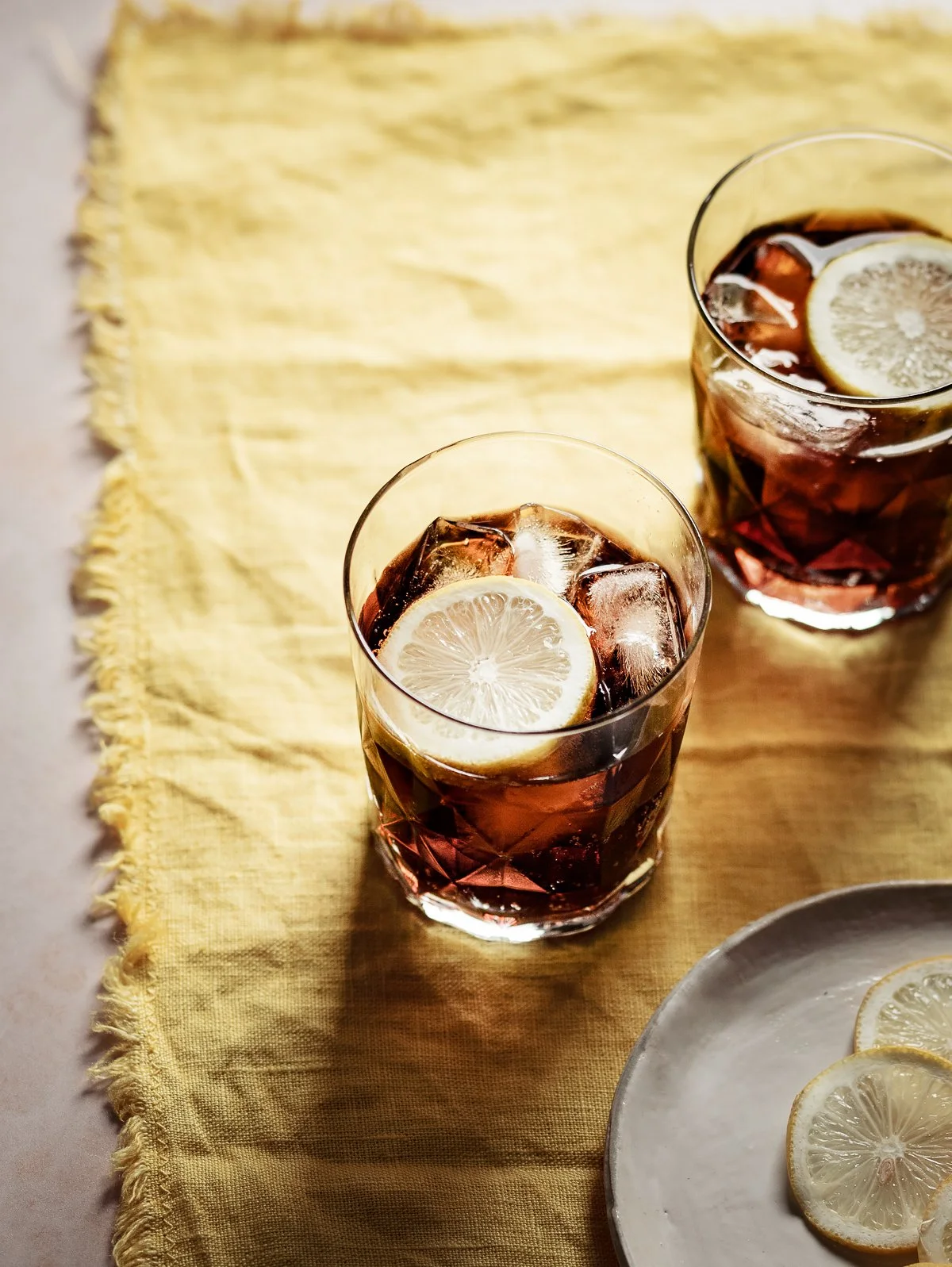 Two glasses of iced tea with lemon slices on a yellow cloth napkin, with a white plate of lemon slices nearby.