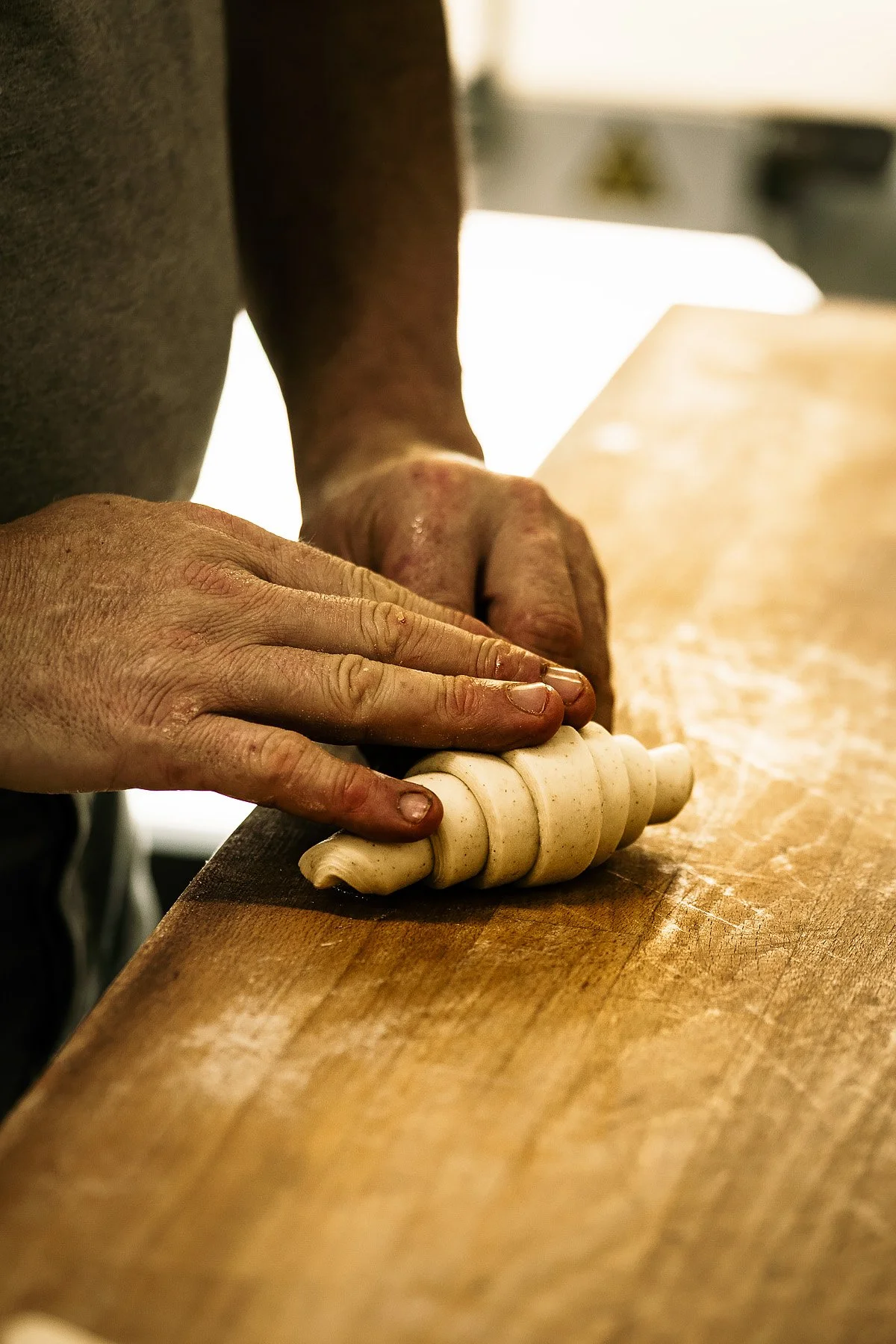 Hands rolling dough into a layered, spiral shape on a wooden surface.