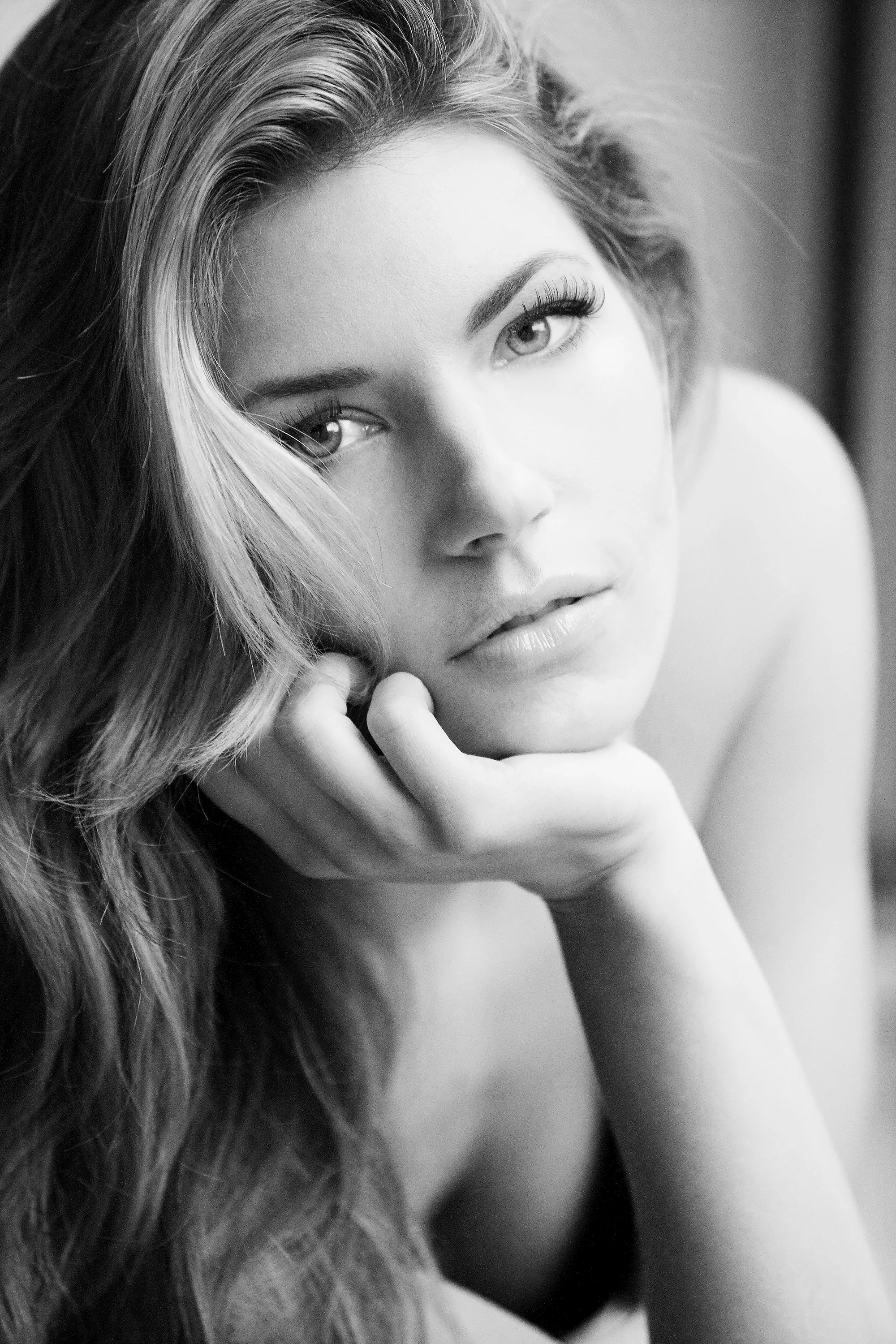 A black-and-white close-up portrait of a woman with long, wavy hair, resting her chin on her hand and looking directly at the camera with an intense expression.
