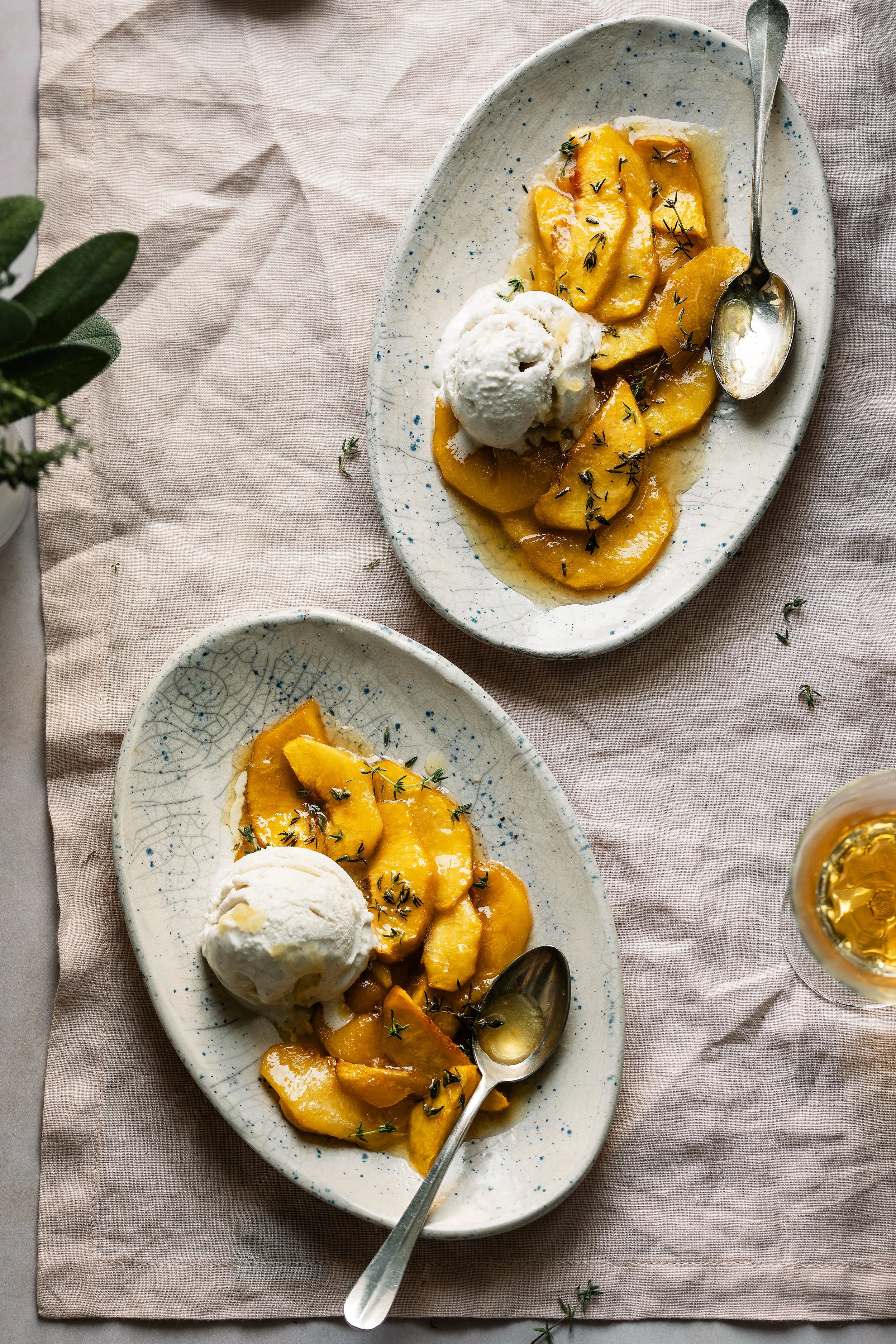 A white oval plate with peach slices topped with herbs and a scoop of vanilla ice cream, next to a silver spoon. A similar dish is partially visible below. The plates are on a light pink fabric tablecloth, with a small plant visible on the left.