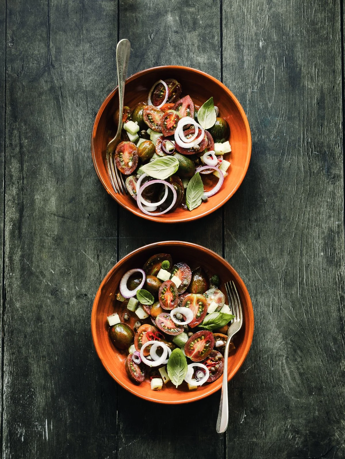 Two orange bowls filled with cherry tomatoes, sliced onions, basil leaves, and green onions on a dark wooden table, with forks placed inside each bowl.