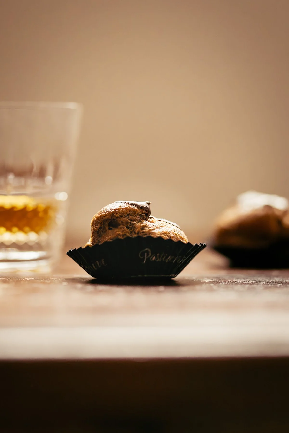 A close-up image of a partially eaten chocolate chip muffin in a black paper cup, with a glass of whiskey and another muffin blurred in the background on a wooden surface.
