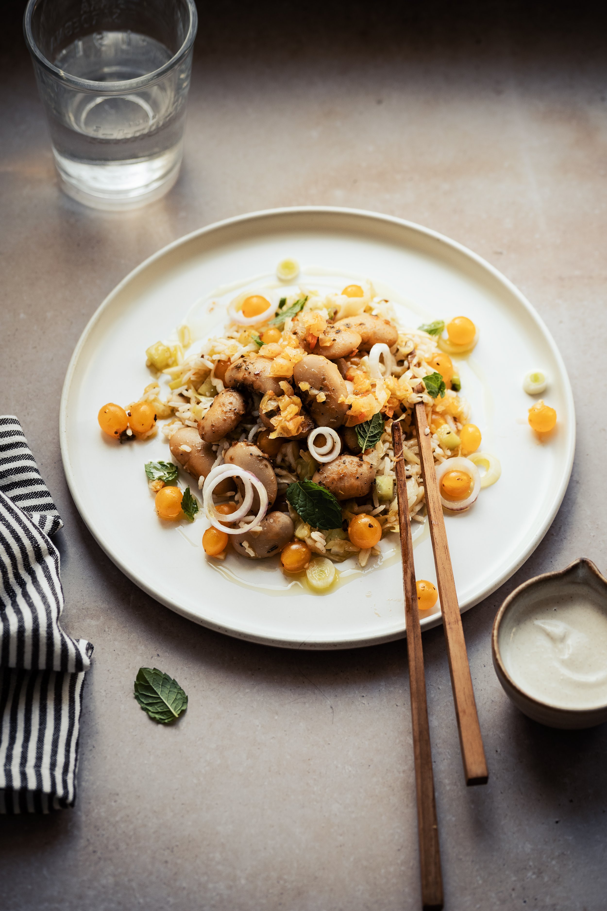 A plate of Asian-style salad with mushrooms, rice noodles, and vegetables, garnished with fresh herbs, with a glass of water and small bowls of sauce nearby on a light-colored surface.