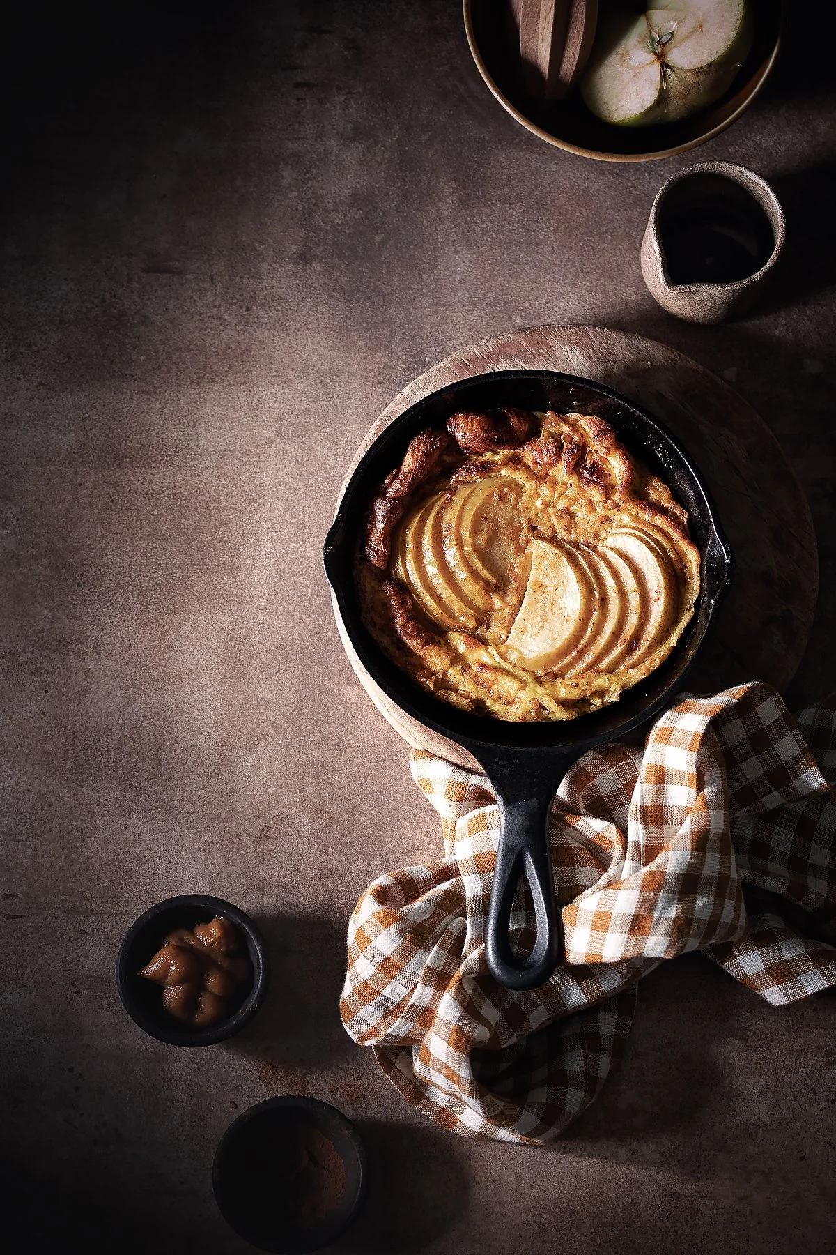 A skillet of apple cobbler topped with sliced apples, placed on a rustic wooden board, with a checkered cloth underneath. A bowl of apples and a small cup of dark syrup are on the table, with a cup of coffee nearby. The setting is dark and cozy.