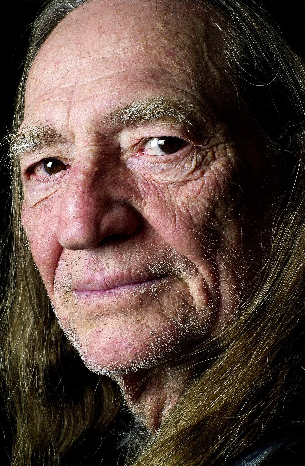 Close-up portrait of an elderly man with long gray hair, wrinkled skin, and a serious expression.