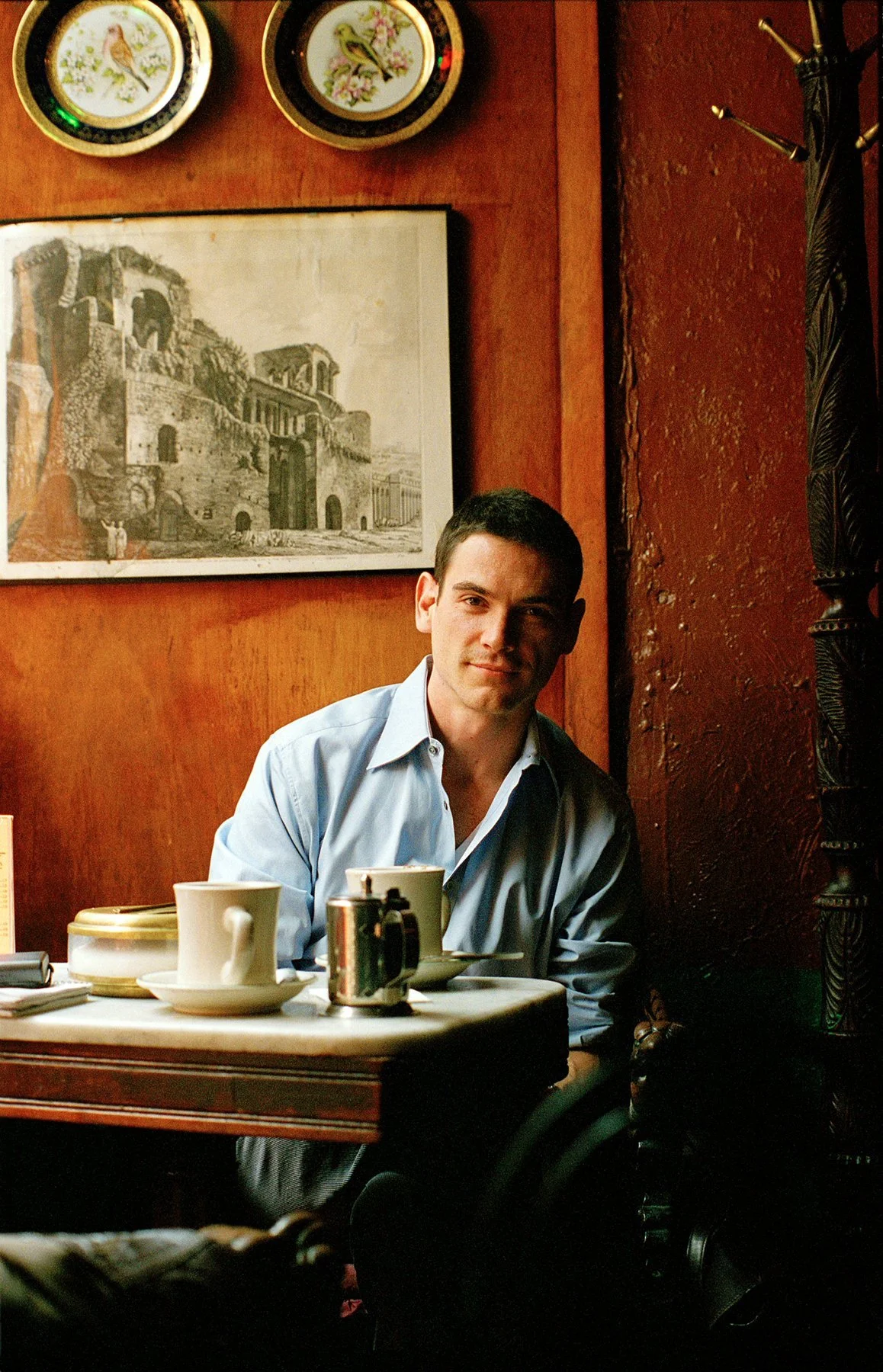 A young man in a light blue shirt sitting at a table in a cozy restaurant. There are cups, a sugar jar, and side plates on the table. The warm reddish-brown wall behind him is decorated with framed artwork of ancient architecture and decorative plate