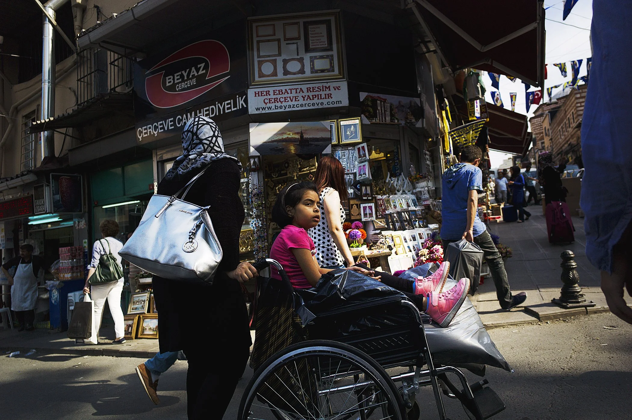 A busy street scene with people walking and shopping. A woman in a black coat with a headscarf and a white purse pushes a stroller carrying a girl in a pink shirt and pink shoes. There is a street vendor stall in the background selling picture frames and other items. Several people are visible in the background, and decorative flags hang across the street.