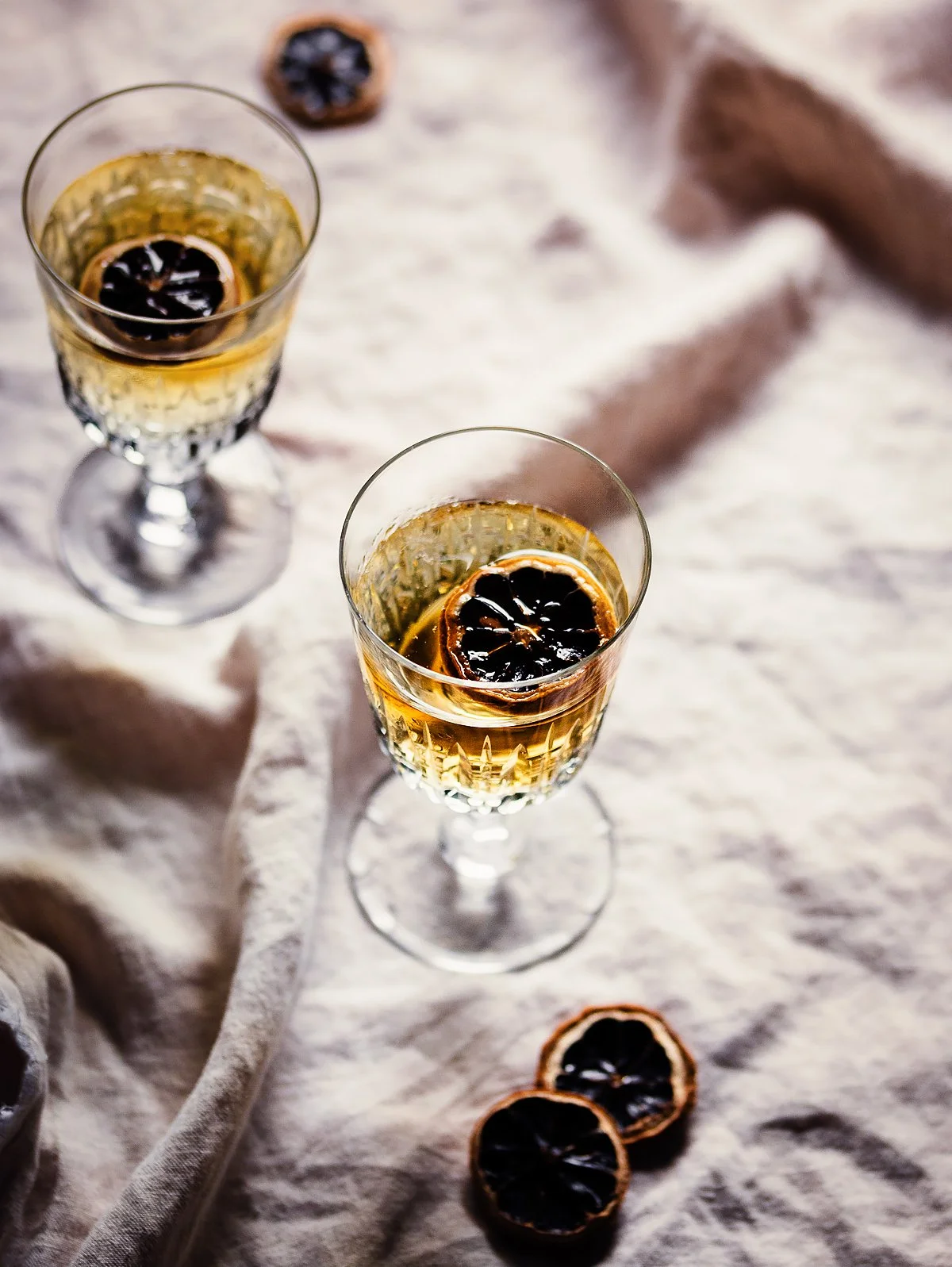 Two elegant champagne flutes filled with sparkling wine, each garnished with a dried orange slice and a black cherry on top, on a textured beige fabric surface with additional dried orange slices nearby.