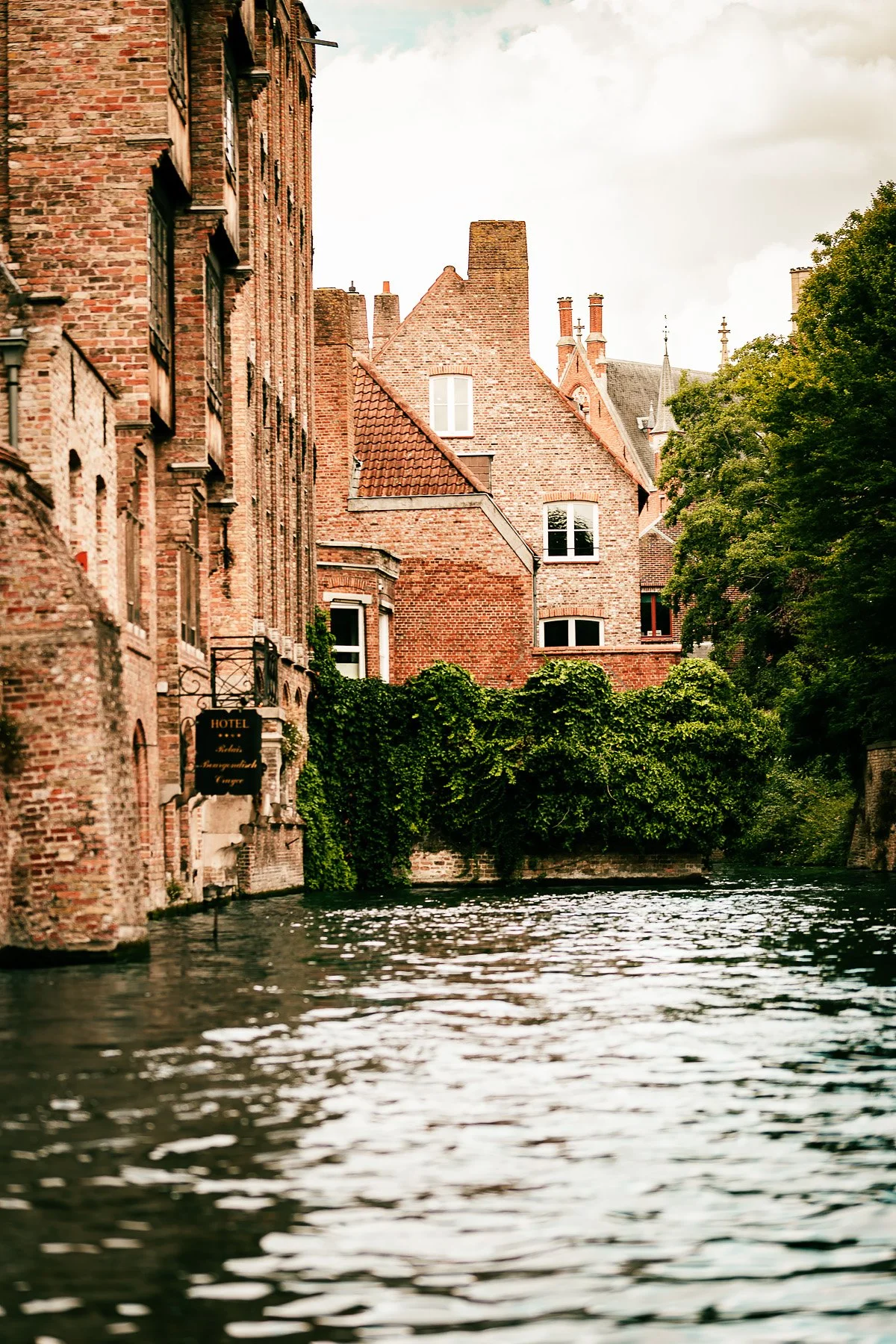 Historic brick buildings with a hotel sign, alongside a canal with rippling water and lush green trees.