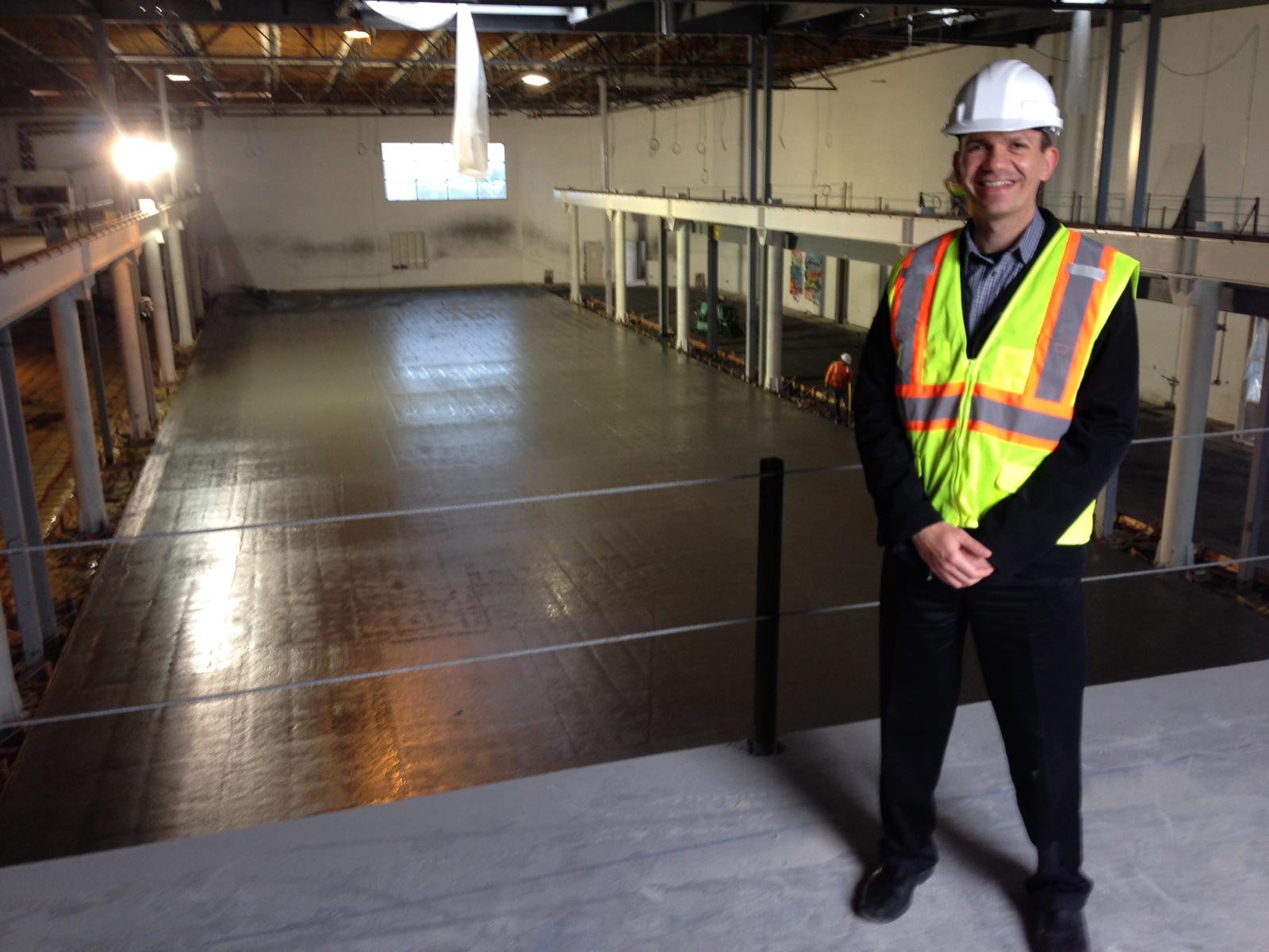 A man wearing a white safety helmet and a bright yellow safety vest with orange reflective stripes, standing inside an industrial or construction area with a concrete floor being prepared for pouring or finishing.