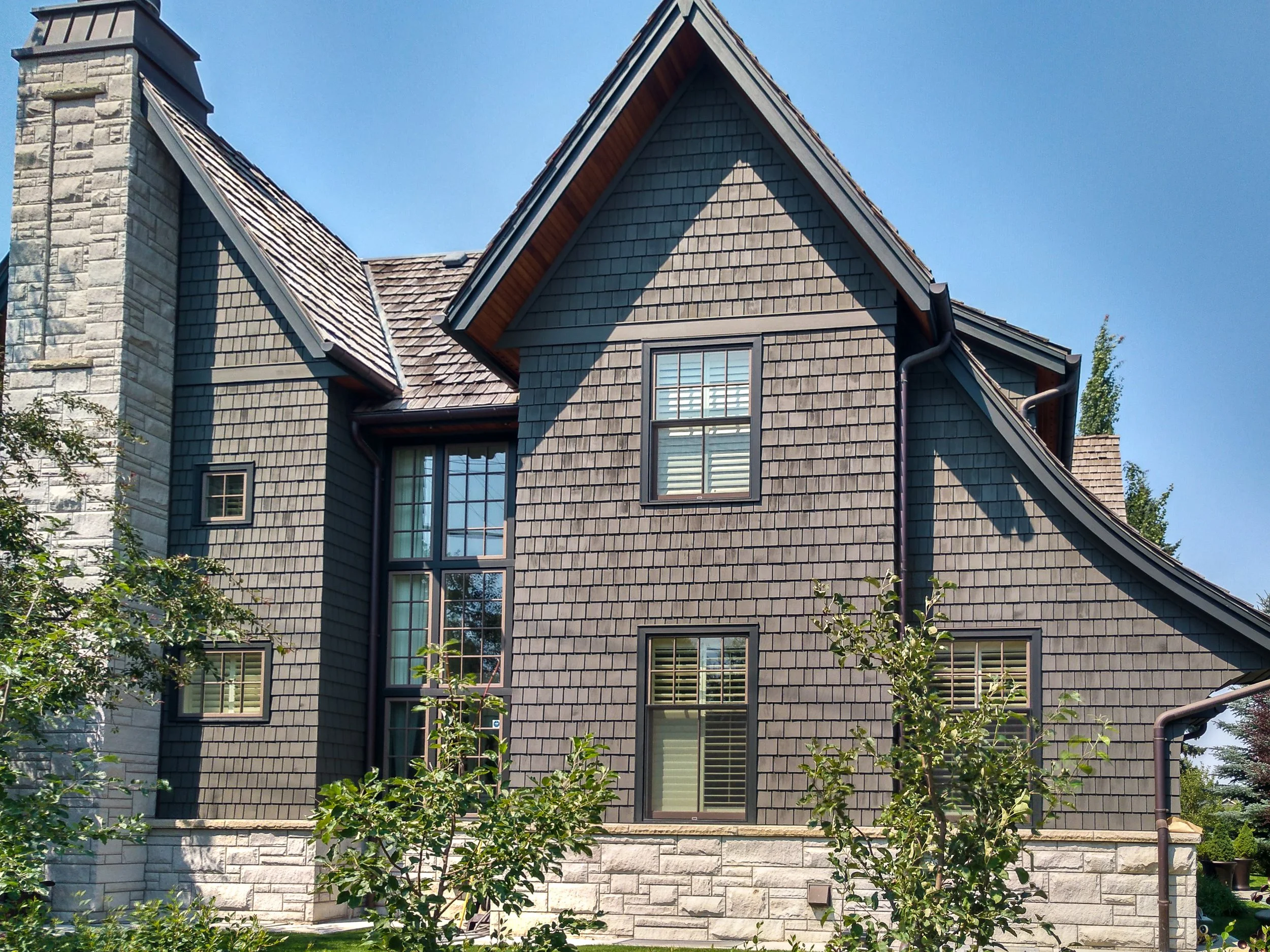 Front view of a house with gray shingles, stone accents, and multiple gabled roofs under a clear blue sky.