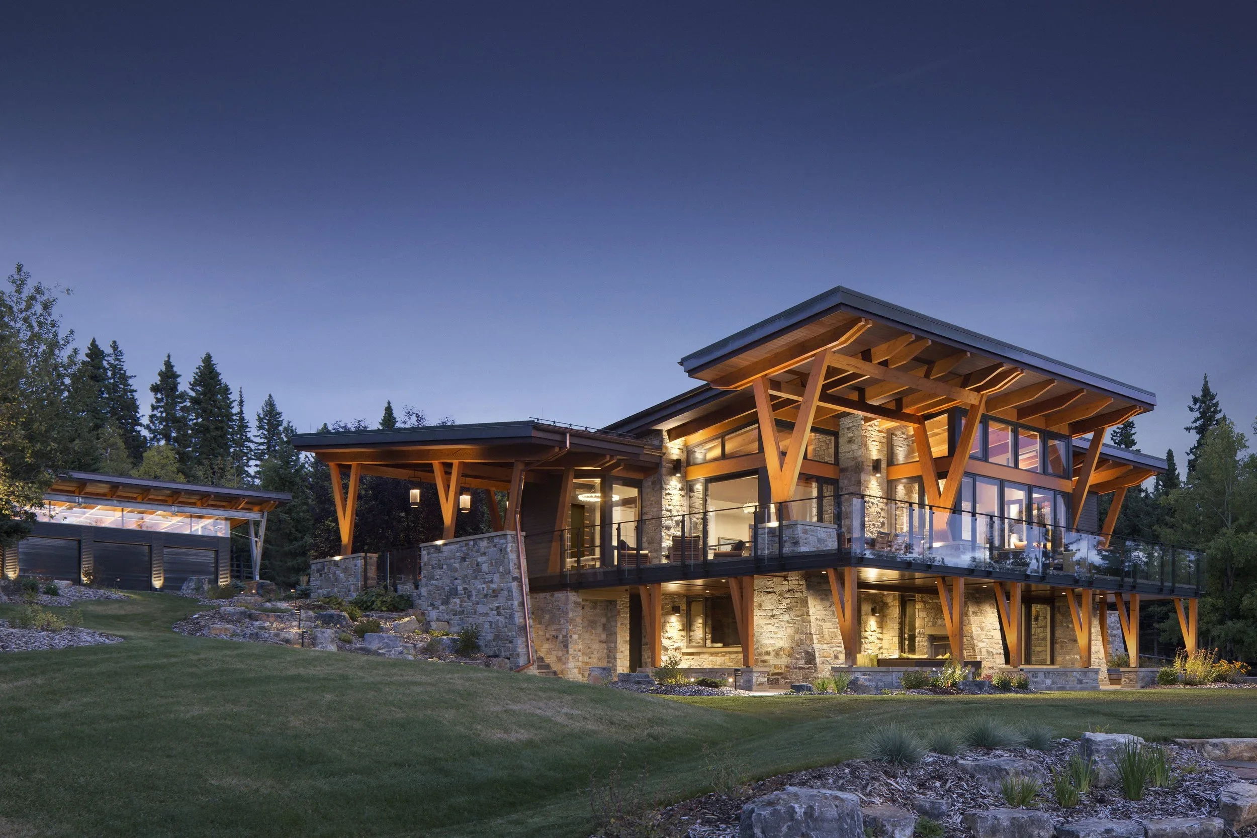 Modern two-story house with large windows, stone and wood exterior, and a spacious balcony, surrounded by a landscaped yard and trees at dusk. Dda Architecture Ltd.