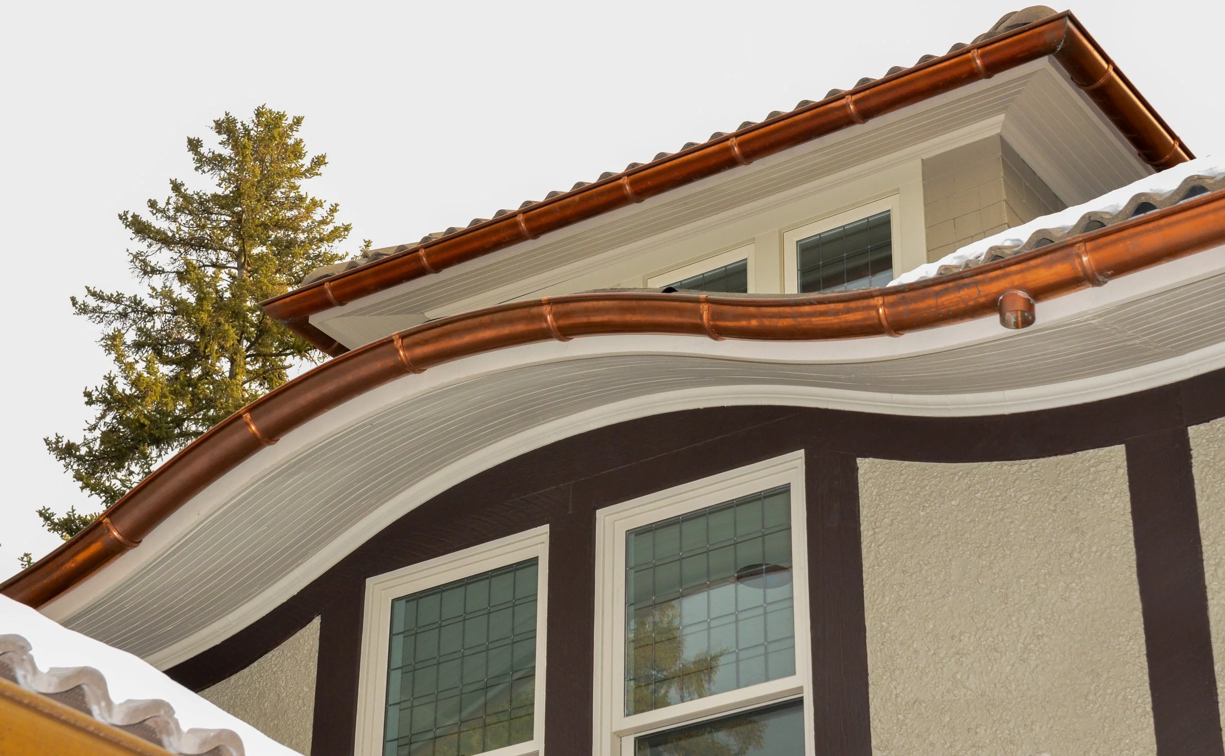 Close-up of a house's roof with copper gutters, upper windows, and a decorative curved edge, with a tall evergreen tree in the background.