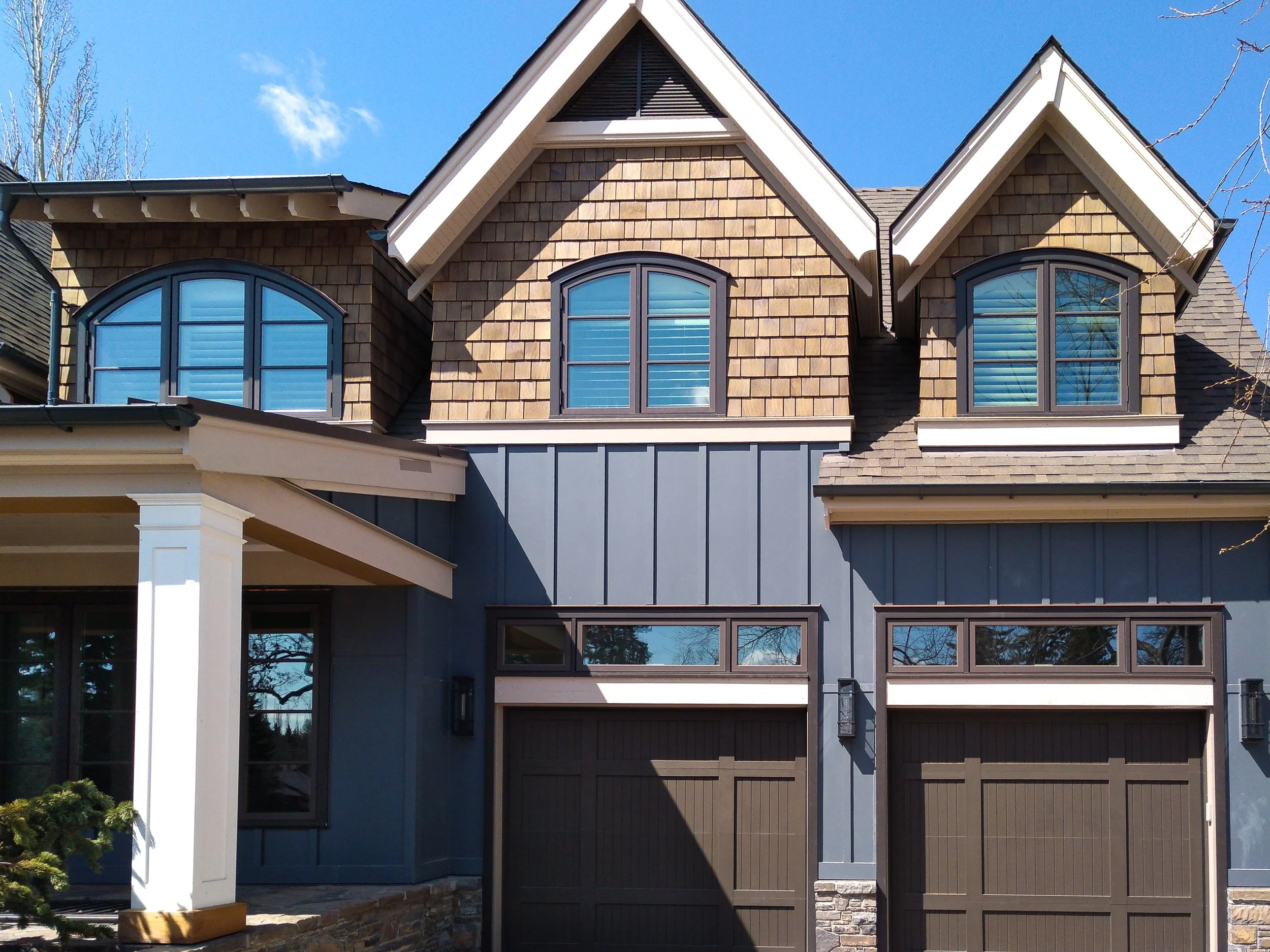 Front view of a two-story house with blue and brown exterior siding, two brown garage doors, and arched windows with shutters under a clear blue sky.