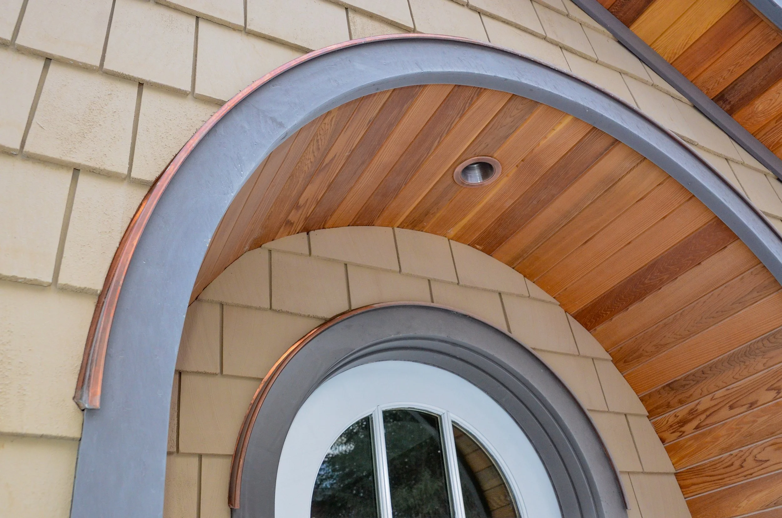 Close-up view of a round window with a metal frame, set in a wall with beige bricks and wooden paneling, with a copper strip around the window for decorative or protective purposes.